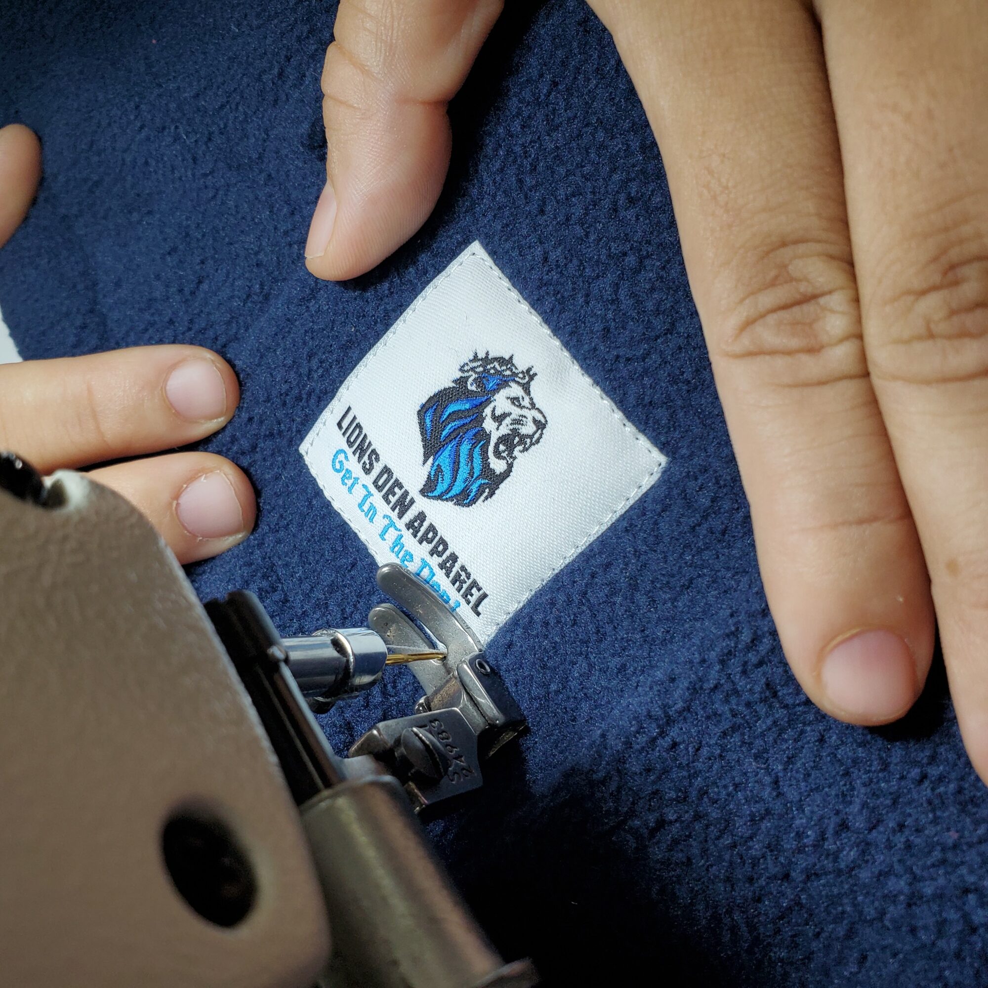 Close-up of a clothing label with a lion logo and text, held by a person's fingers on a blue fabric background.