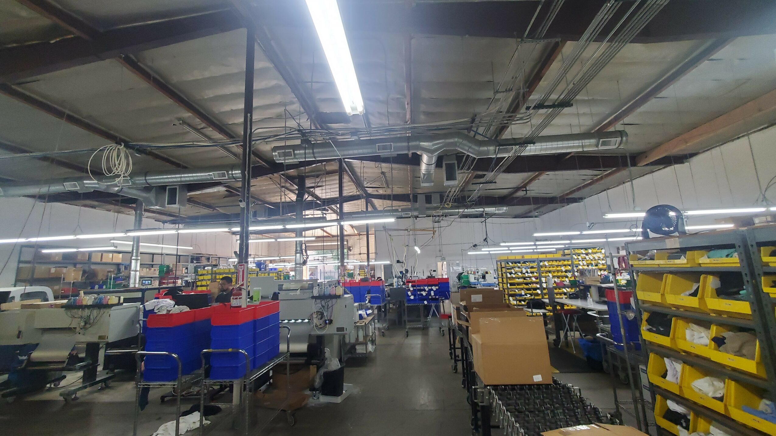 Interior of a warehouse or factory with shelves, carts, and equipment, illuminated by fluorescent lights, ceiling pipes visible.