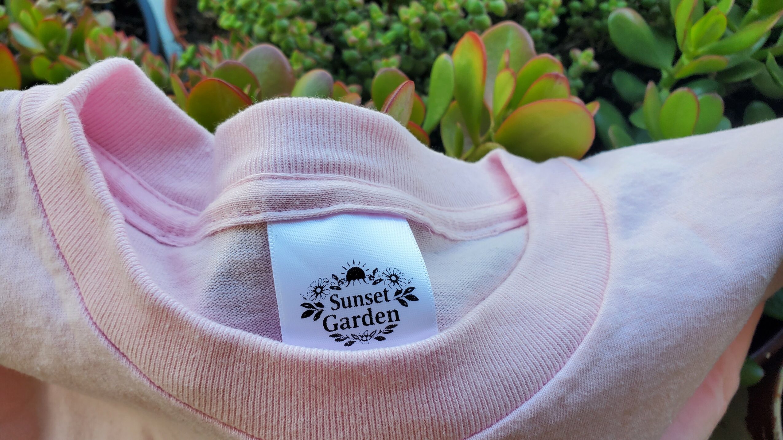 Close-up of a pink and white shirt collar with a label reading 'Sunset Garden' against green plants.