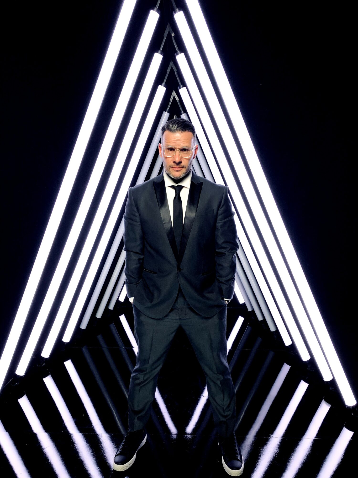 Man in a suit standing in front of illuminated geometric light installation, facing forward, hands in pockets.