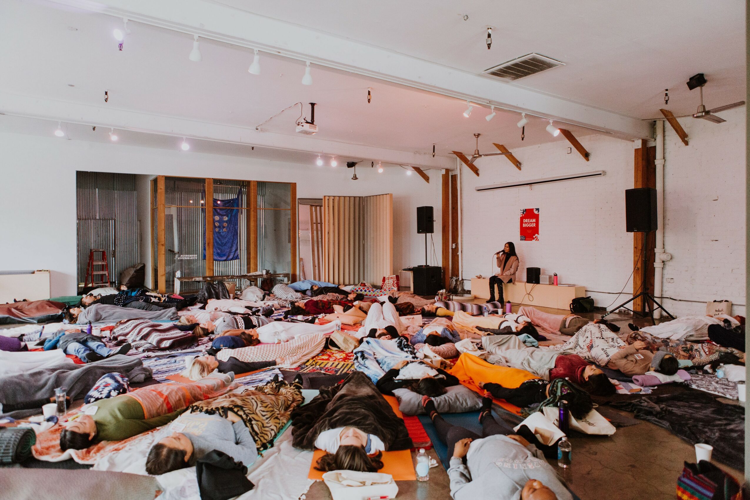 Large group of people lying on the floor in a room with white walls and wooden floor, some with eyes closed.