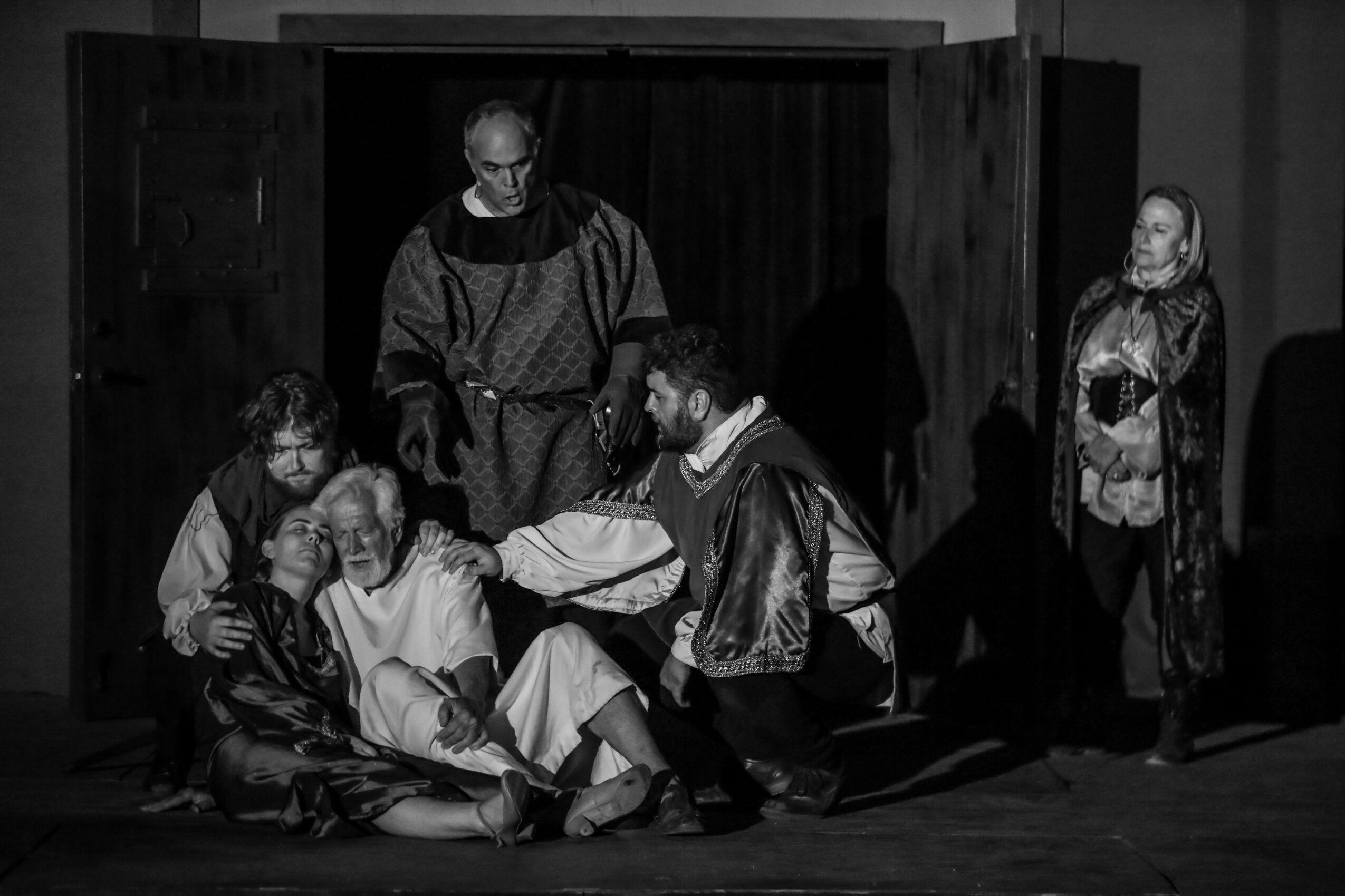 Group of six people on stage, some sitting and some standing, engaged in a scene, with a dark background.