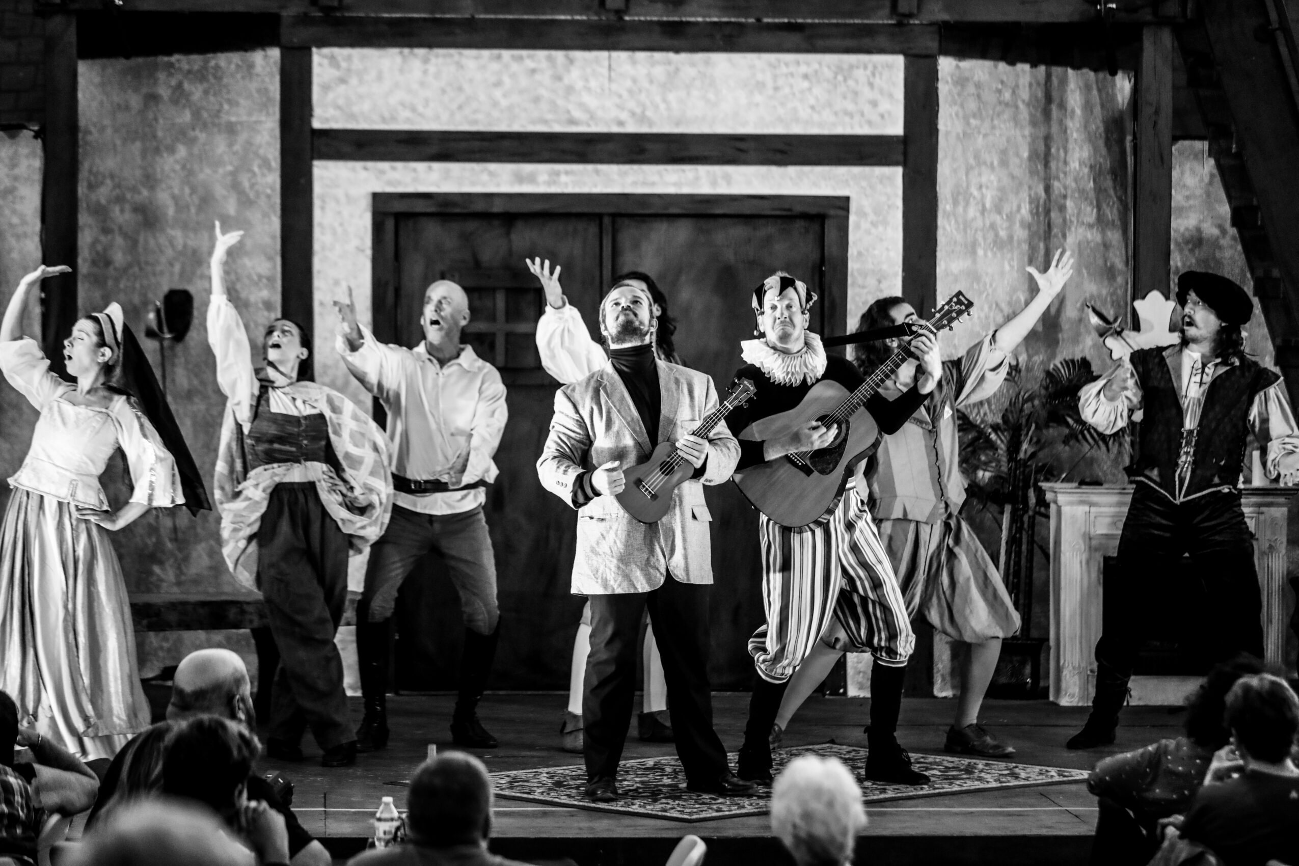 Group of performers on stage, some playing guitars, others dancing, in a theatrical setting with an audience watching.