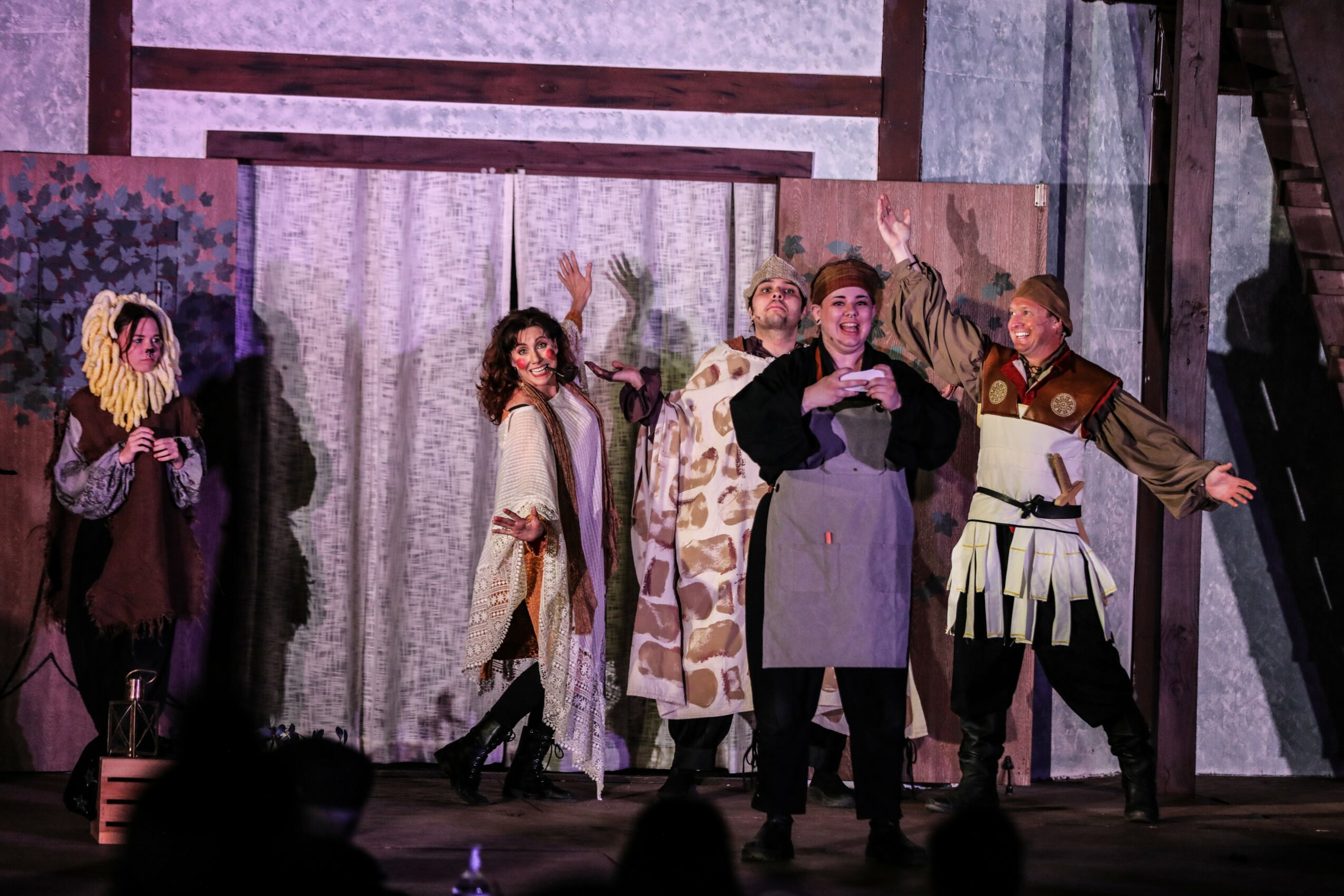 Group of actors performing on stage with costumes and set pieces, some gesturing and smiling.