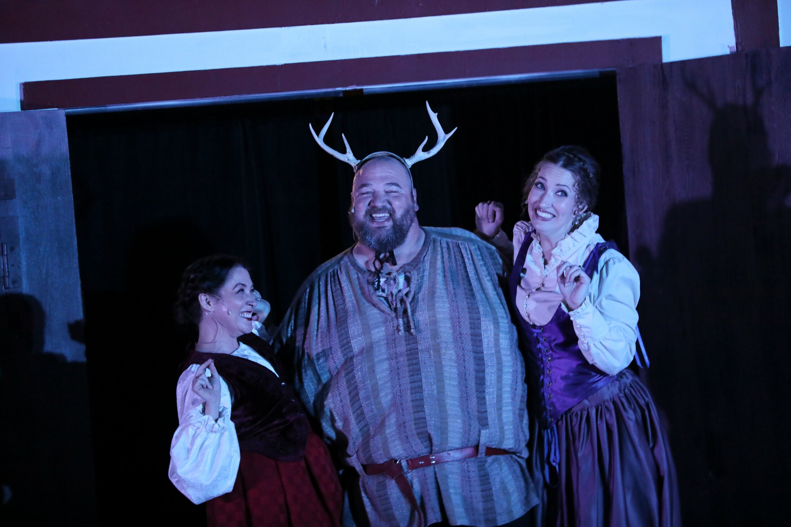 Three people on stage, one with antlers, smiling and posing, with a dark background and wooden panels.