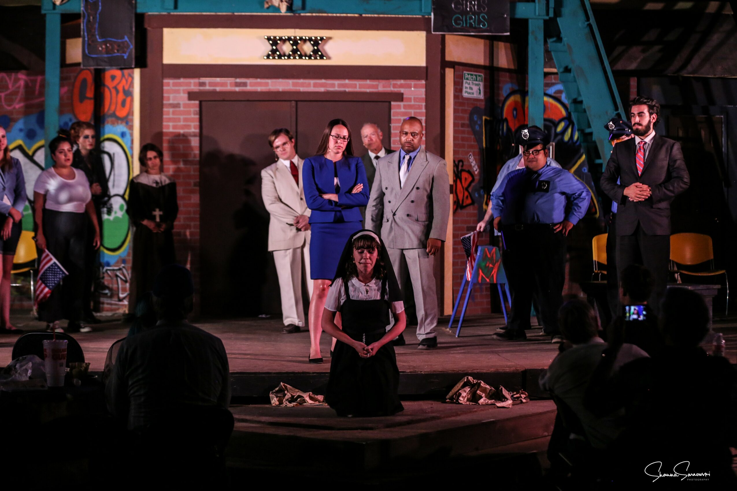 Group of people on stage, some standing and one kneeling in front, with colorful graffiti background and audience in foreground.