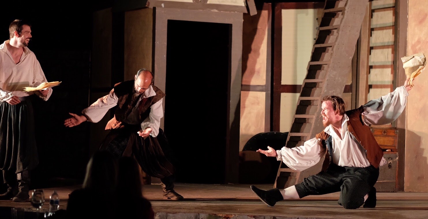Four actors perform on stage, two standing and two kneeling, with a set resembling a rustic building in the background.