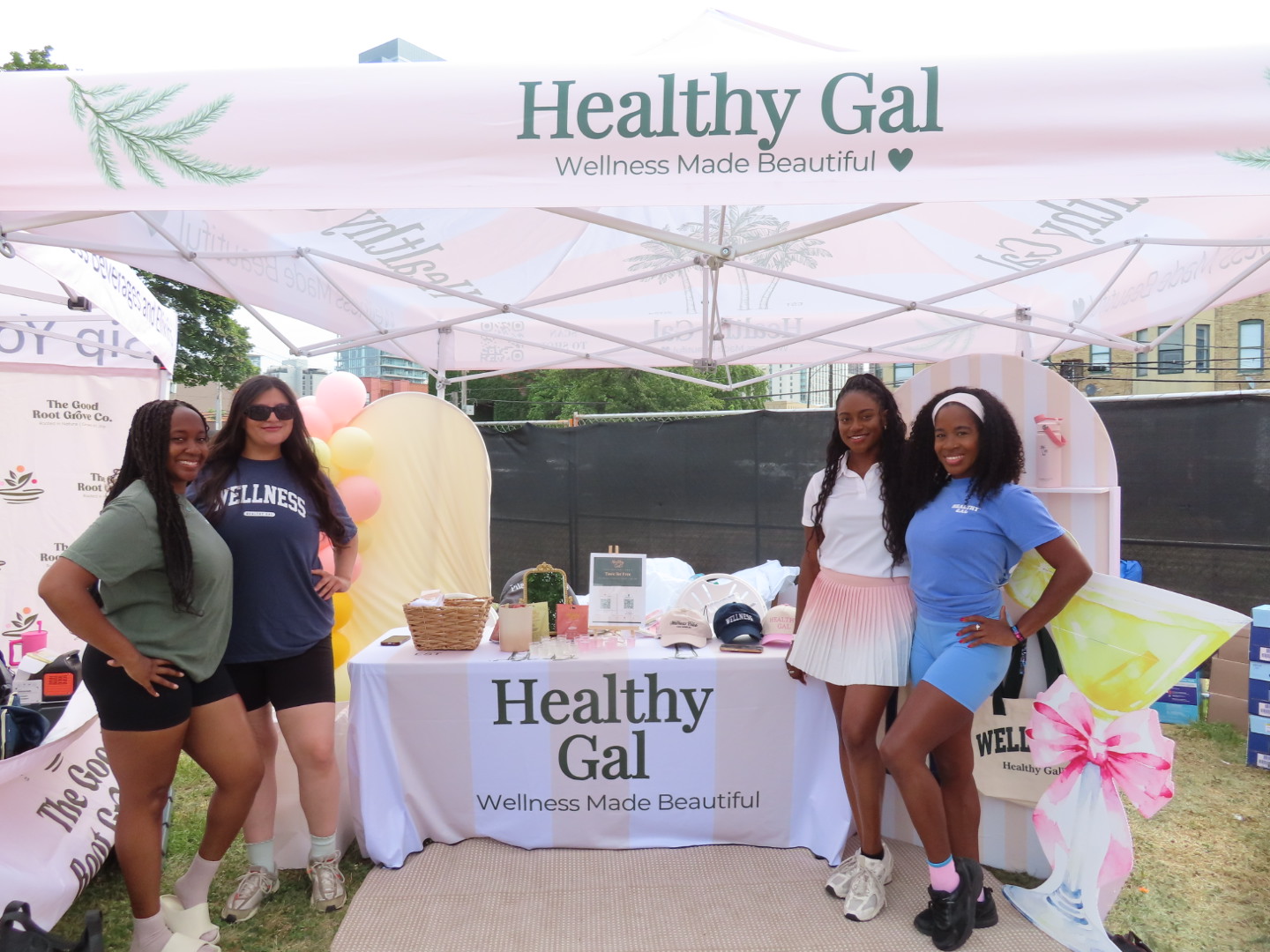 Four women standing behind a table at a health awareness event outdoors, with a banner reading 'Healthy Gal'.