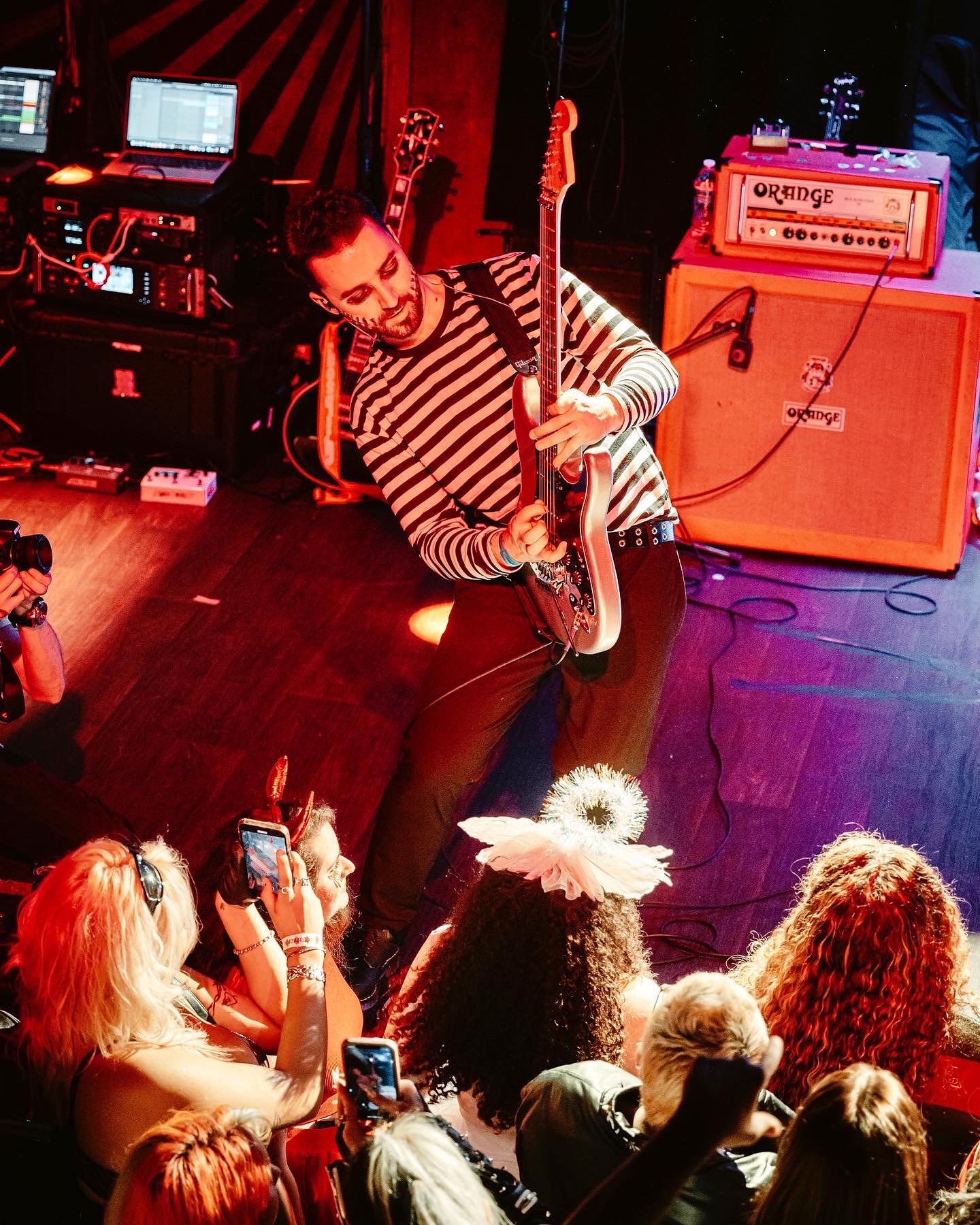 Person playing electric guitar on stage with musical equipment and audience taking photos.