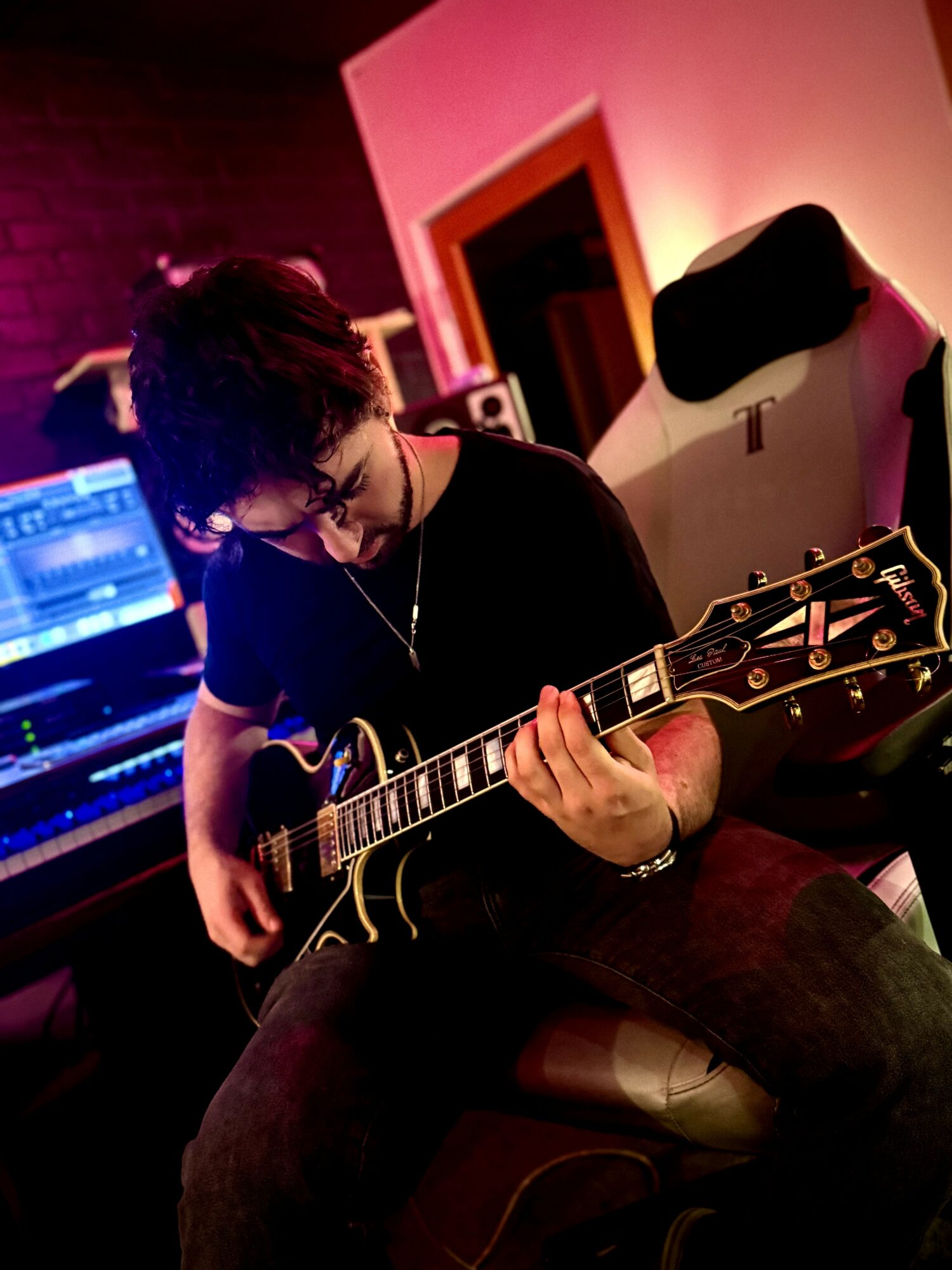 Person playing an electric guitar in a music studio with colorful lighting, wearing glasses and a black shirt.