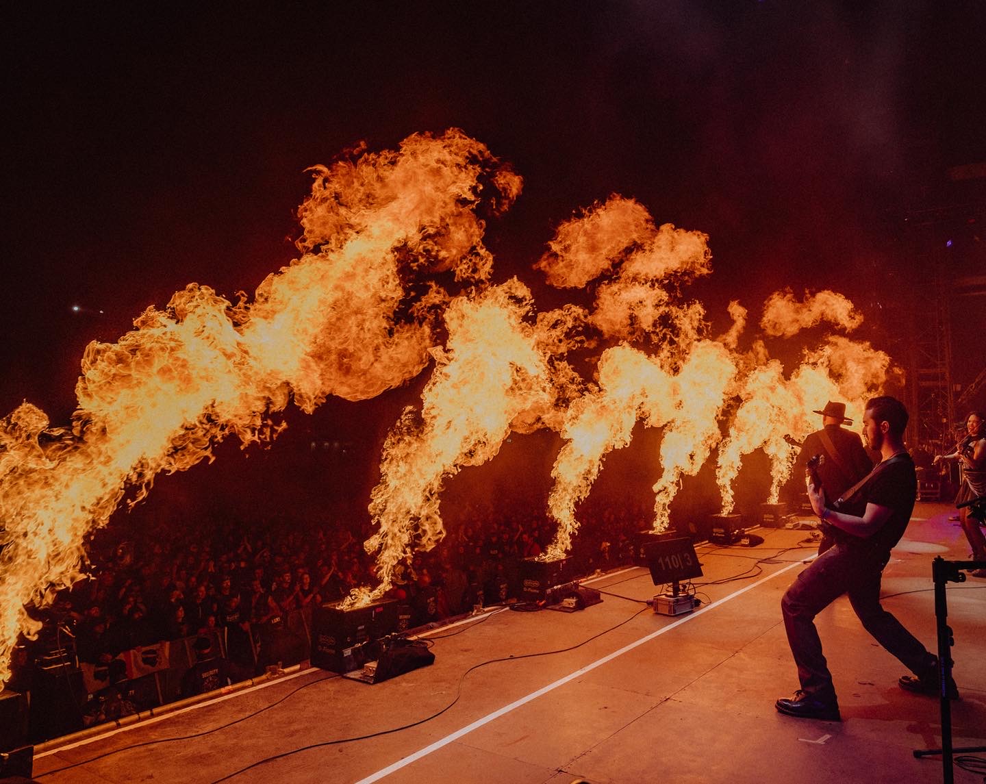 Performer on stage with fire effects creating large flames, audience visible in the background, person taking photos, dark setting.