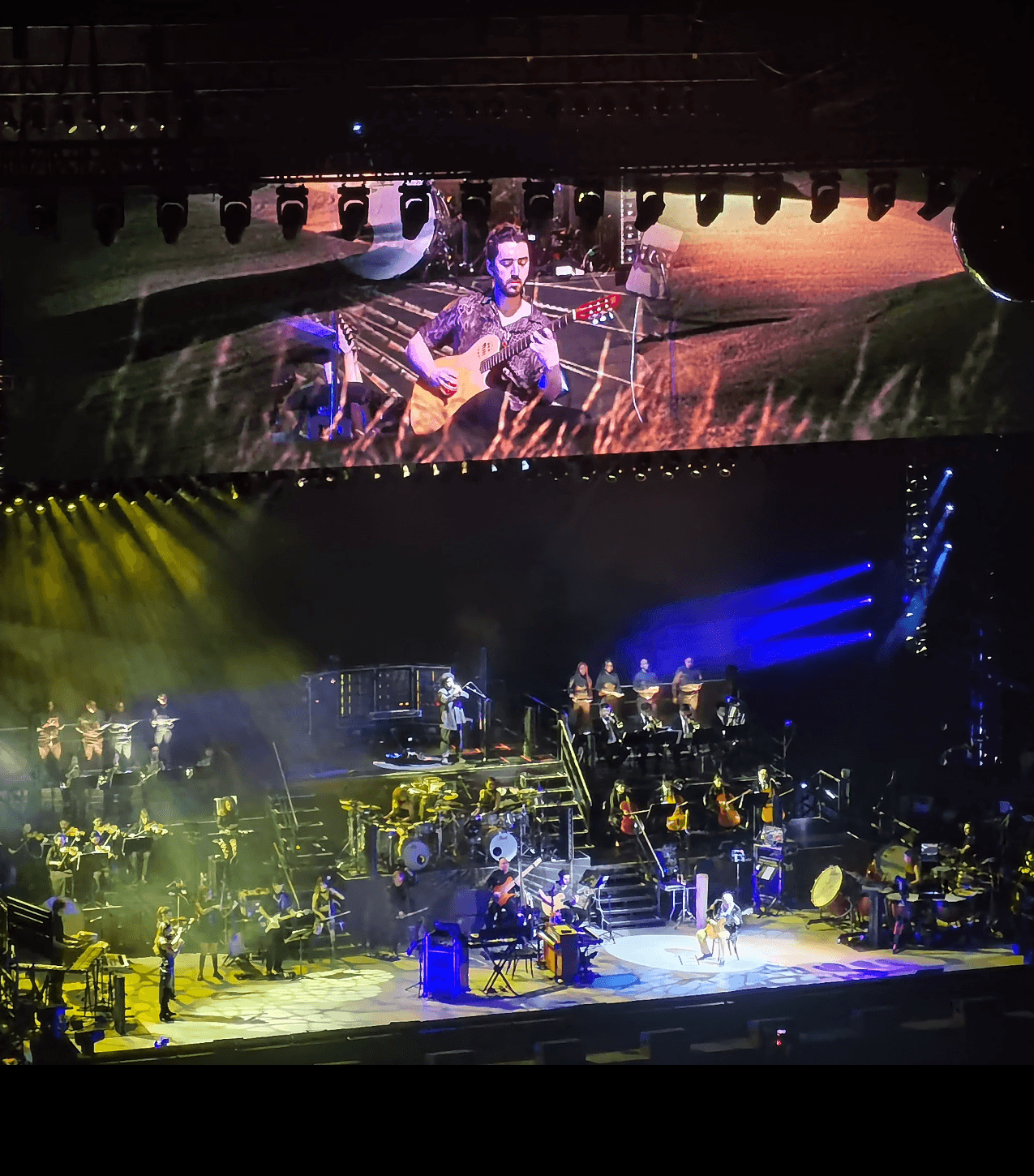 Concert stage with musicians, large screen showing a guitarist, colorful lighting, and stage equipment.