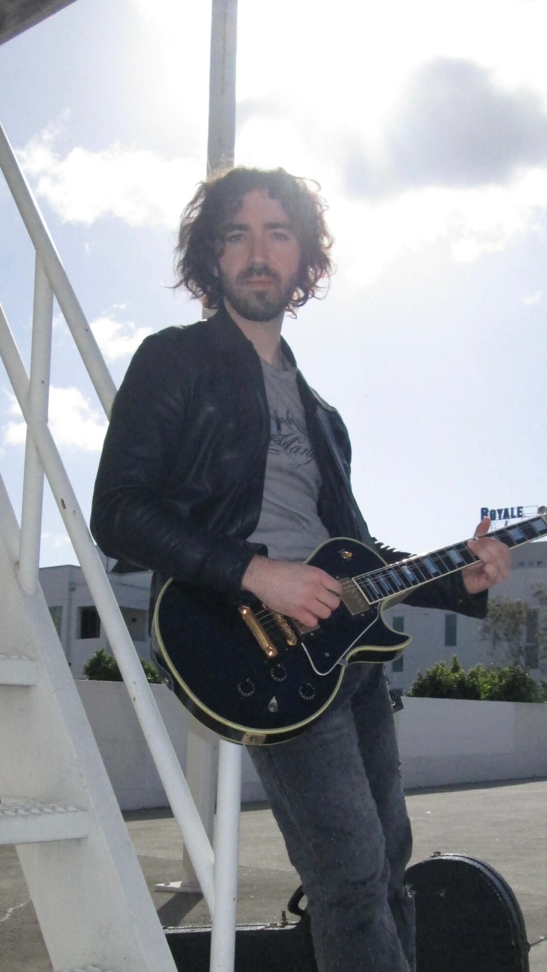 Person with curly hair playing a black electric guitar outdoors, standing near a white staircase, with a cloudy sky background.
