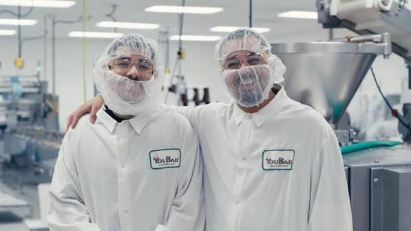 Two people in protective clothing and hairnets in a factory or laboratory setting, smiling and standing close together.