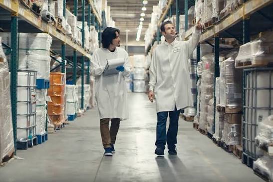 Two scientists in lab coats walking through a warehouse aisle with shelves of supplies, discussing inventory or research materials.