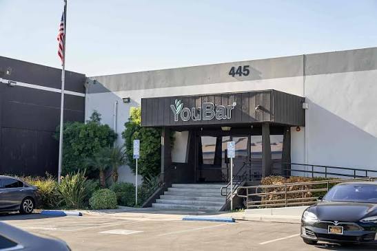 Building with a sign that reads 'YouBar' and the number 445, with stairs leading to the entrance, trees, and parked cars.