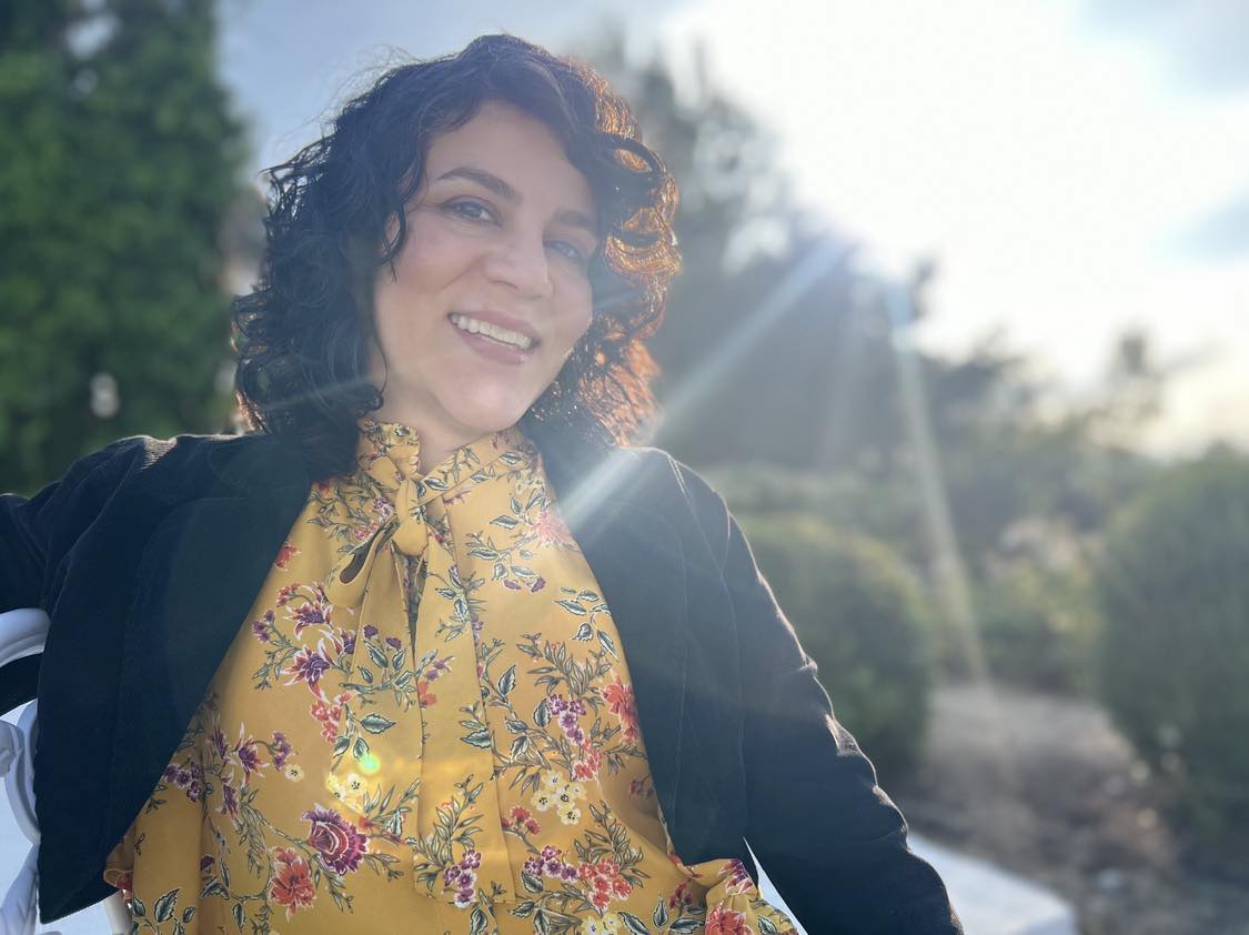 Smiling woman with curly hair outdoors, sunlight shining, wearing a yellow floral blouse and black jacket.