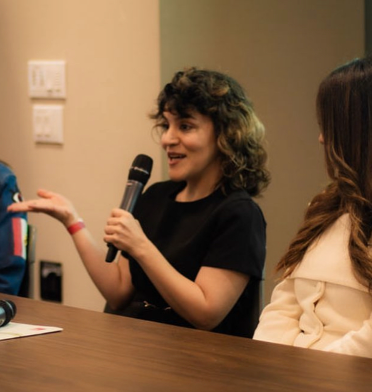 Woman speaking into a microphone, gesturing with her hand, seated at a table with two other people nearby.