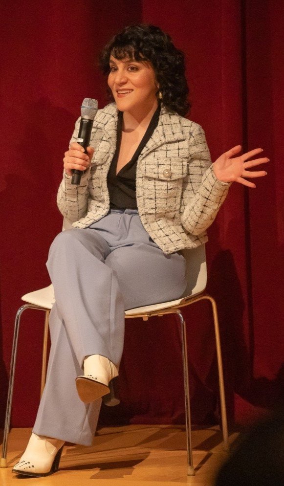 Woman sitting on a chair holding a microphone, gesturing with her other hand, in front of a red curtain.