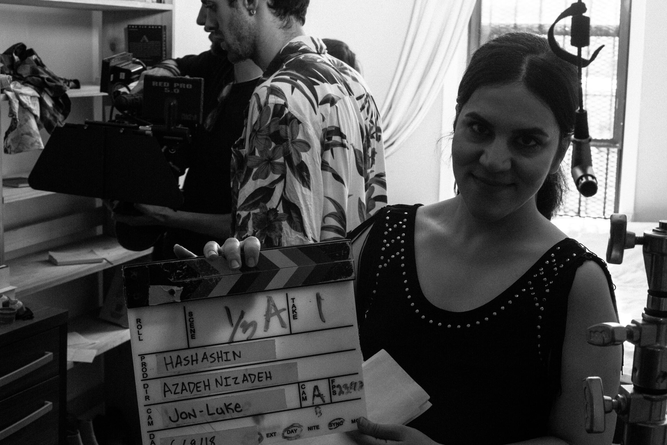 Two people in a room, one woman smiling at the camera, another person working with equipment in the background, black and white photo.