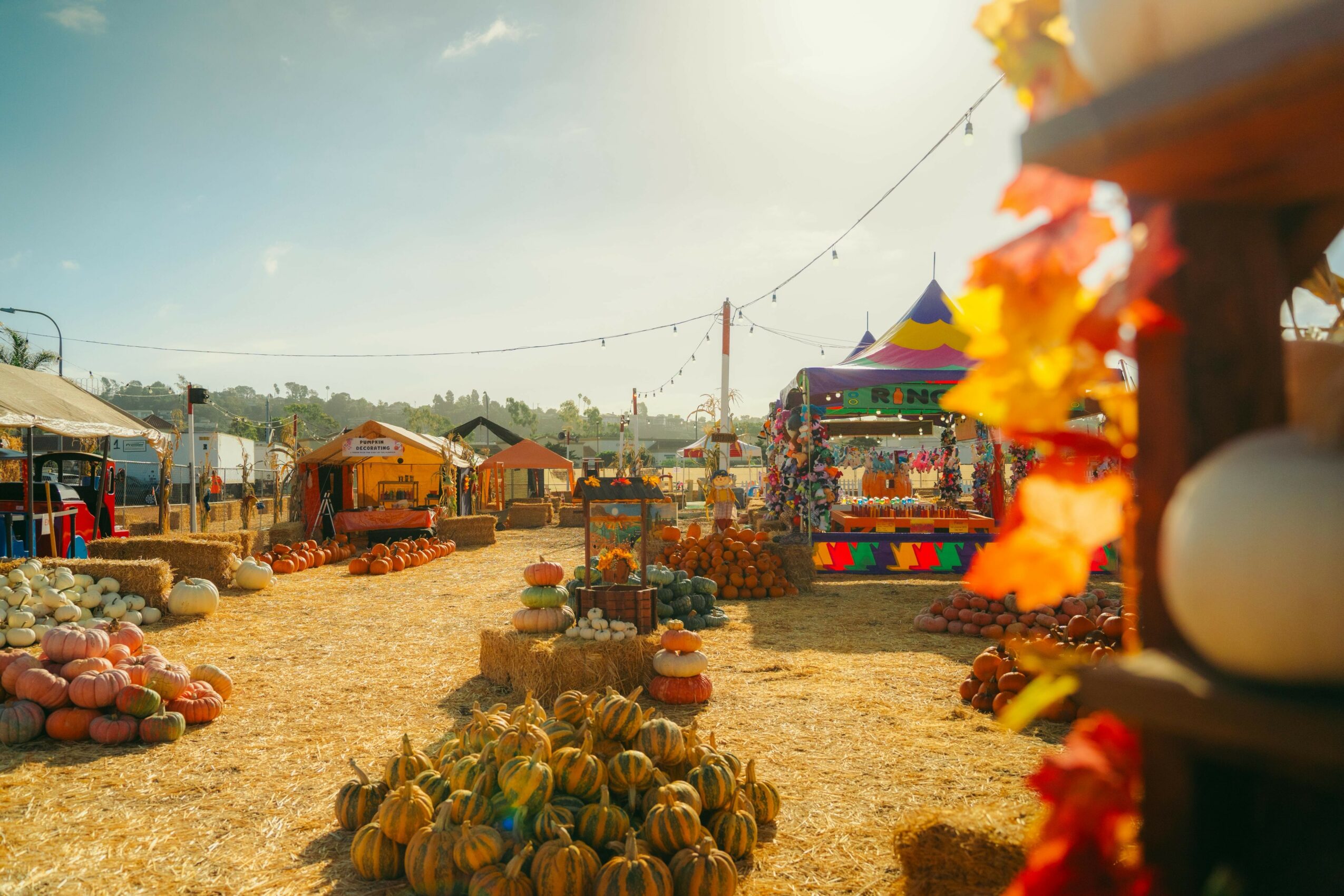 Outdoor market with stalls, pumpkins, and colorful decorations under a clear sky, with a carnival ride in the background.