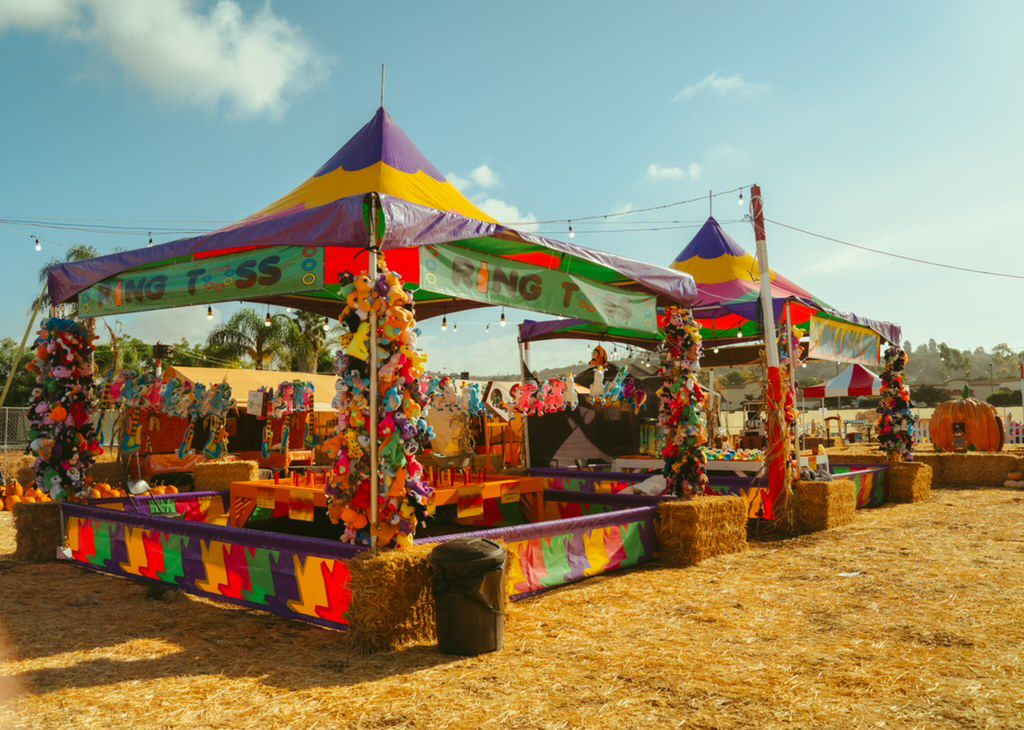 Colorful carnival game booths with tents, decorations, and prizes on a sunny day, set on dry grass.