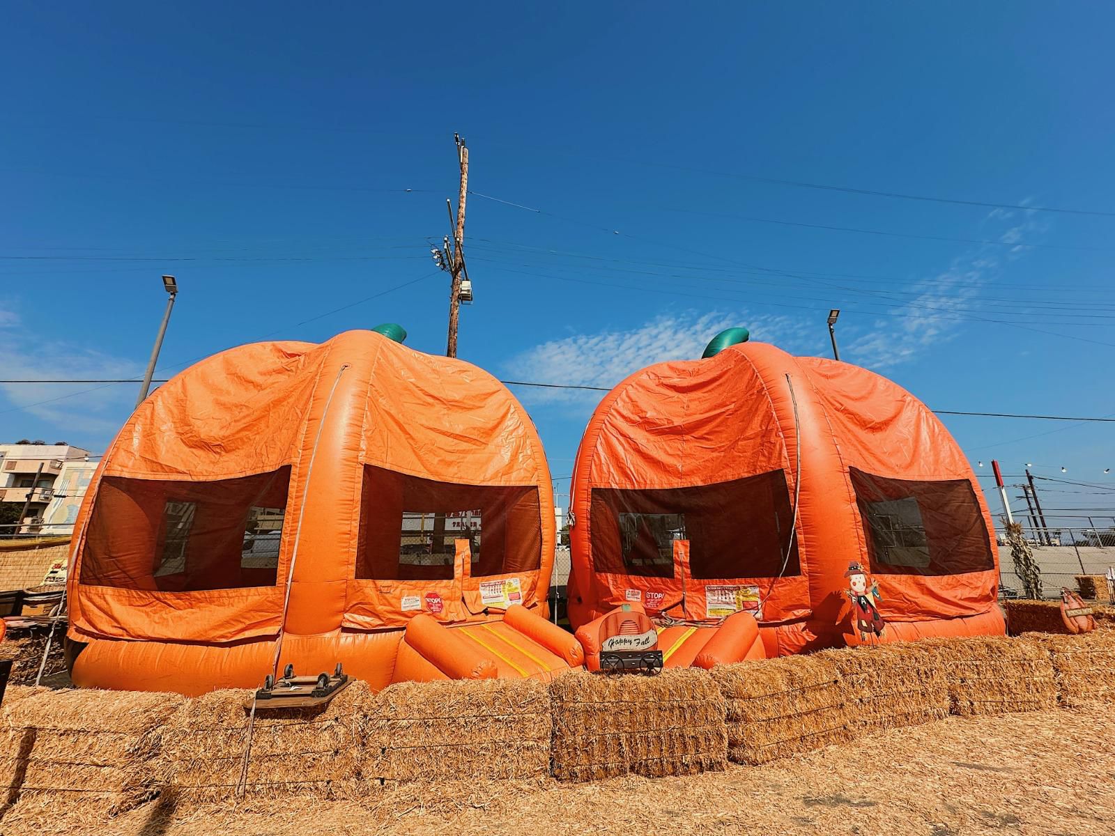 Two large orange inflatable structures resembling pumpkins with black windows, set on a dirt ground under a blue sky.
