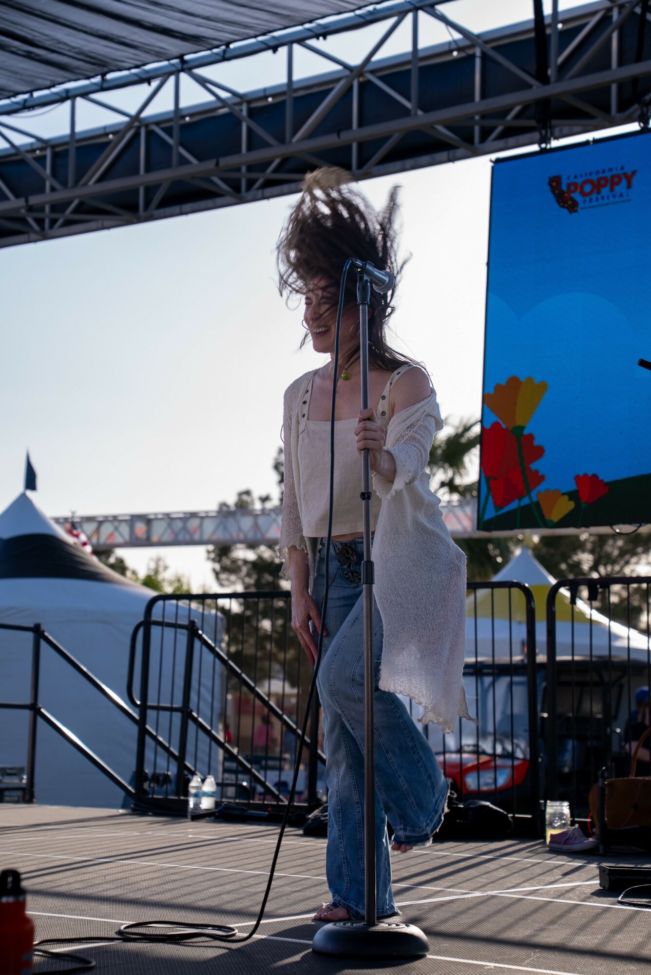 Person with messy hair singing into a microphone on stage outdoors, with a large screen behind and a metal railing in front.