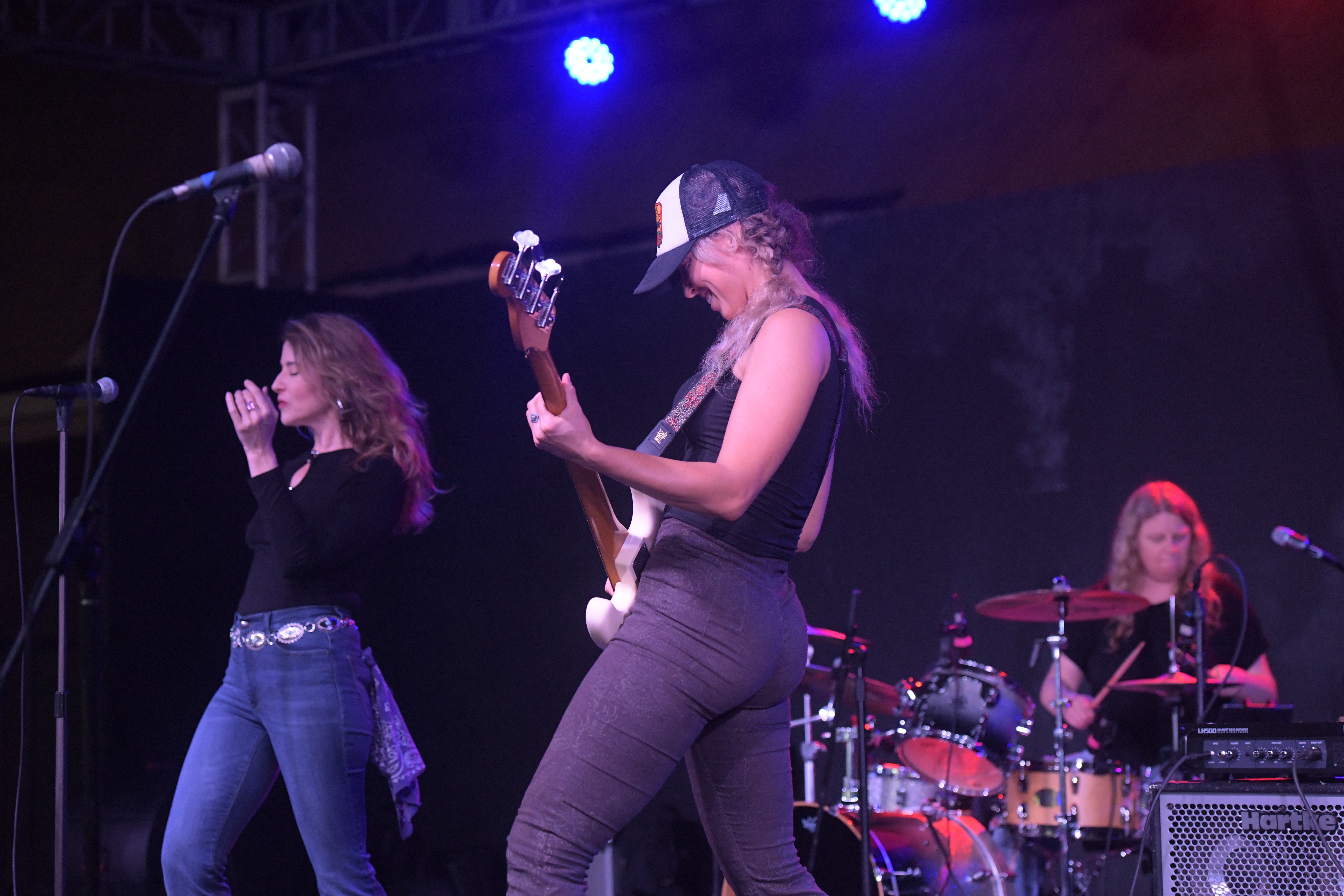 Two women performing on stage with a drummer in the background, one singing and the other playing guitar, stage lighting visible.