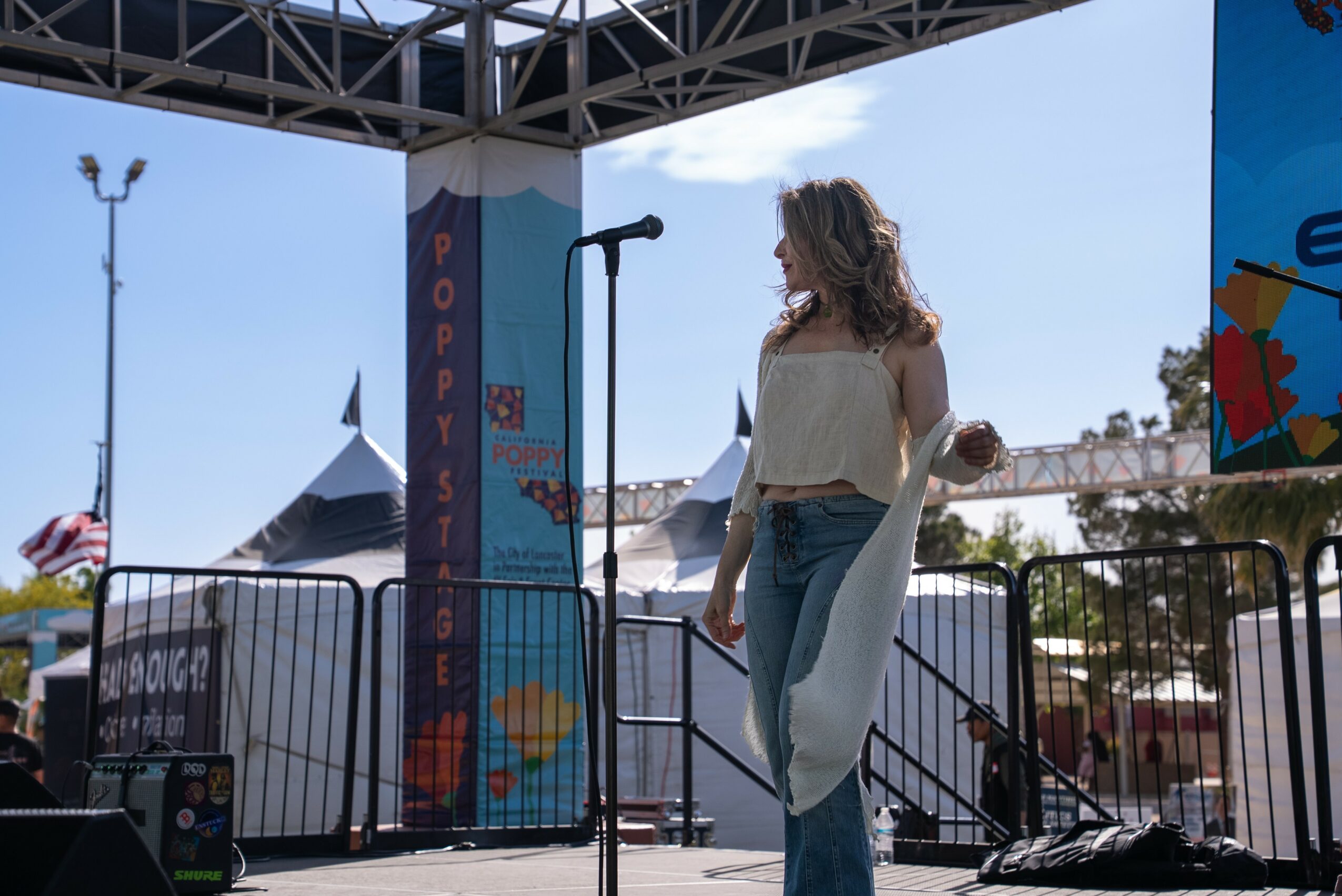 Woman standing on stage with microphone, outdoors, tents and flags in background, sunny day.