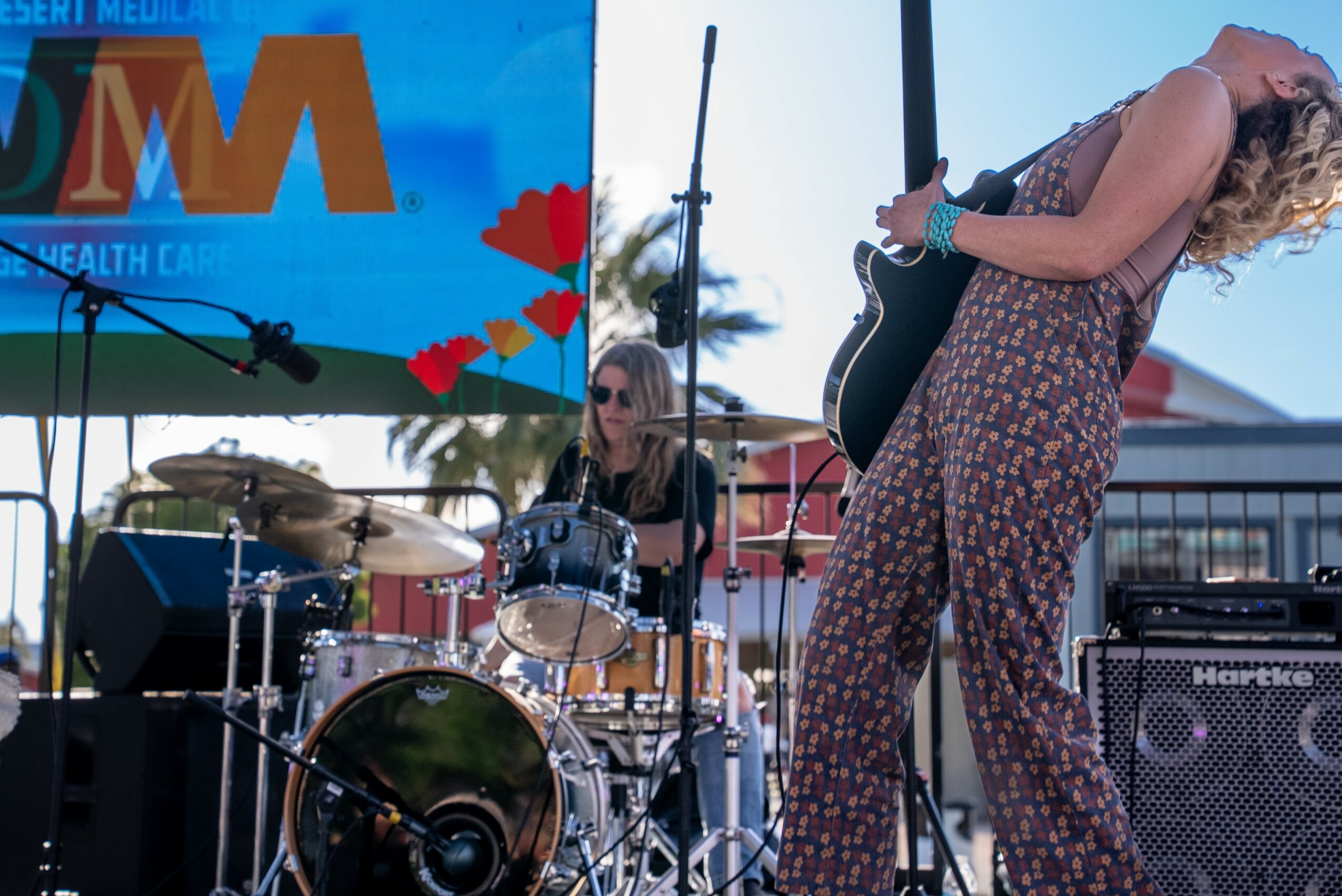 Two women perform on an outdoor stage, one playing drums and the other singing with a guitar, with a large screen behind them.
