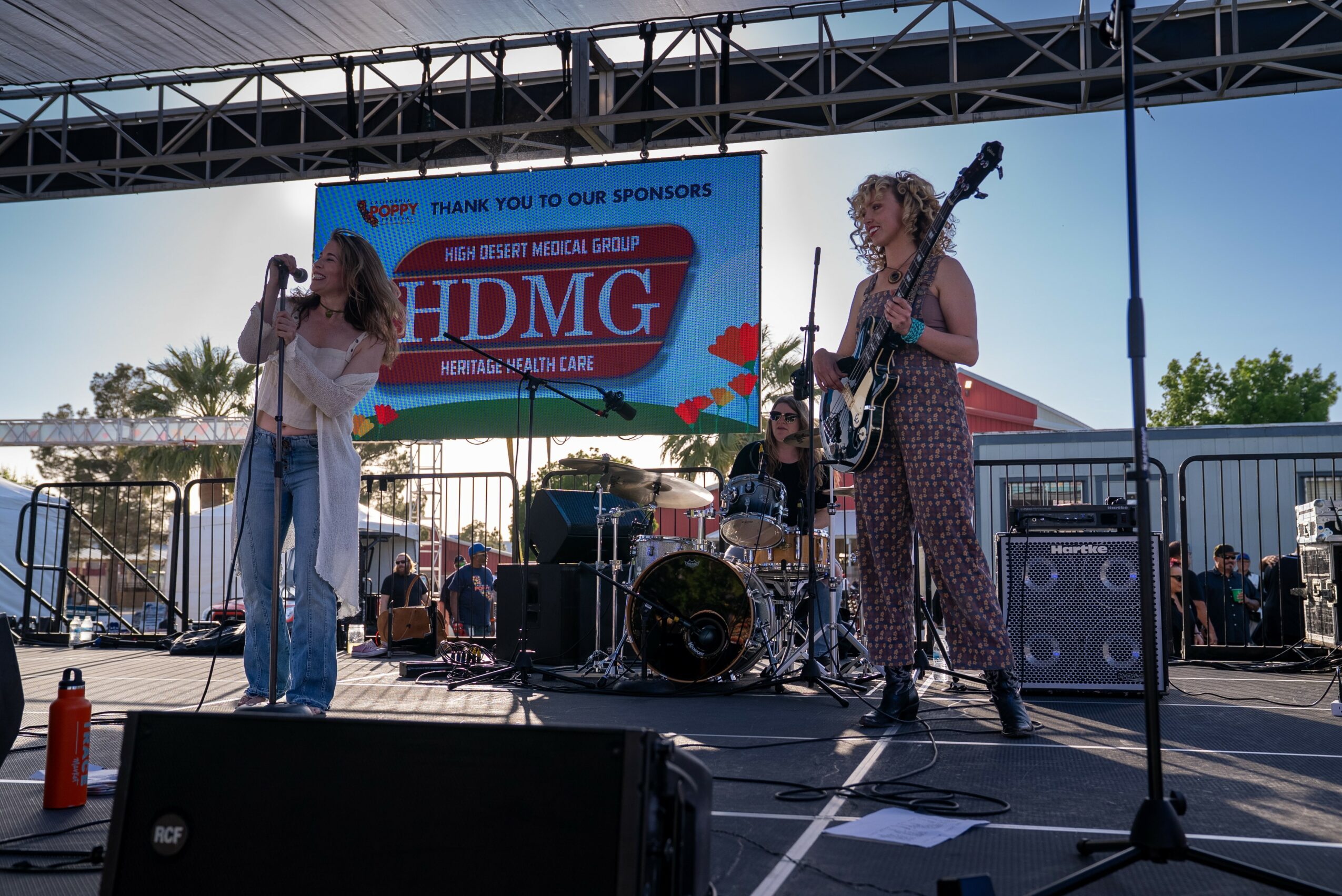 Two women perform on an outdoor stage with a drum set and microphone, a large screen behind them displays 'HDMG' and event details.