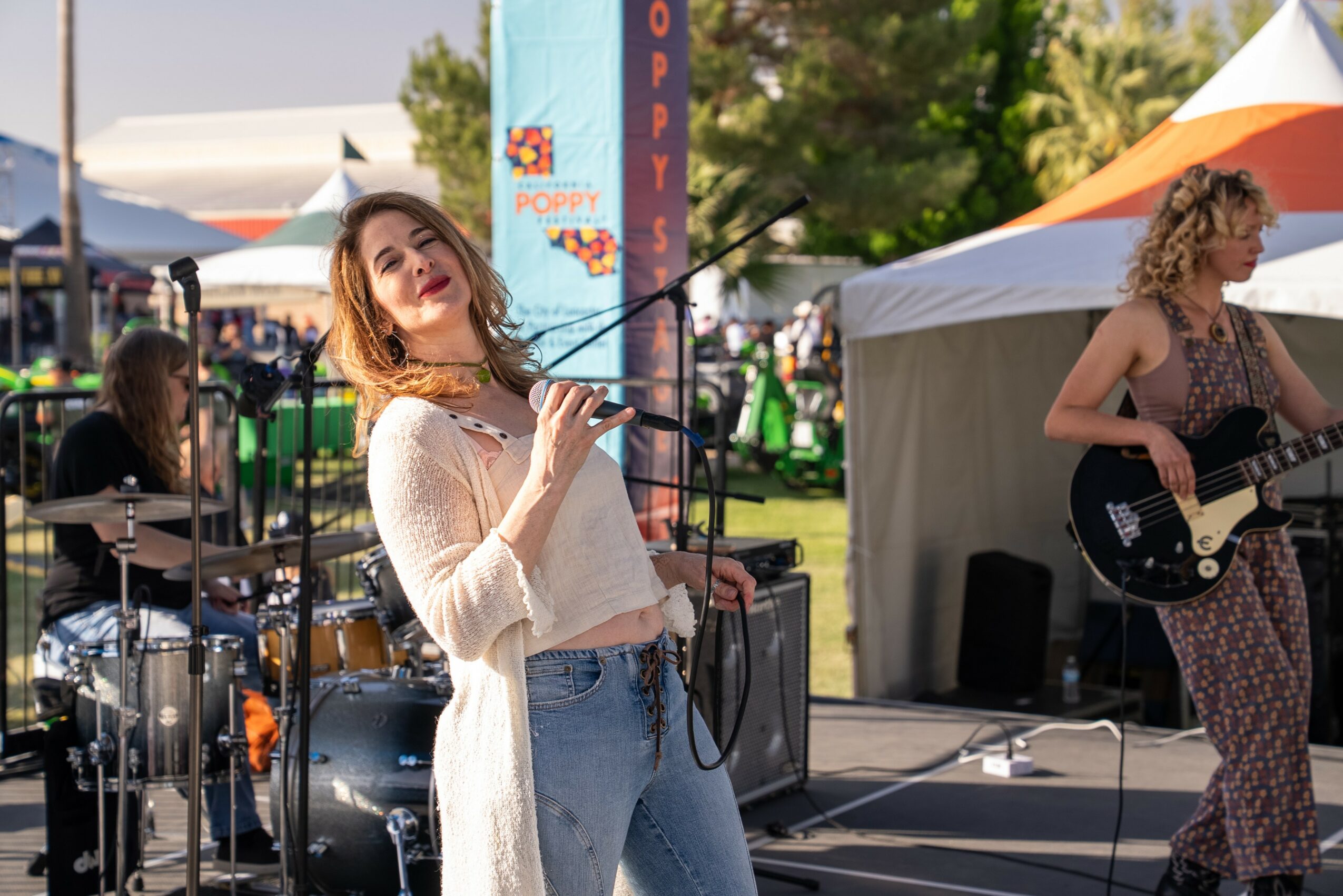 Three women performing music outdoors with tents and trees in the background, one singing and two playing instruments.