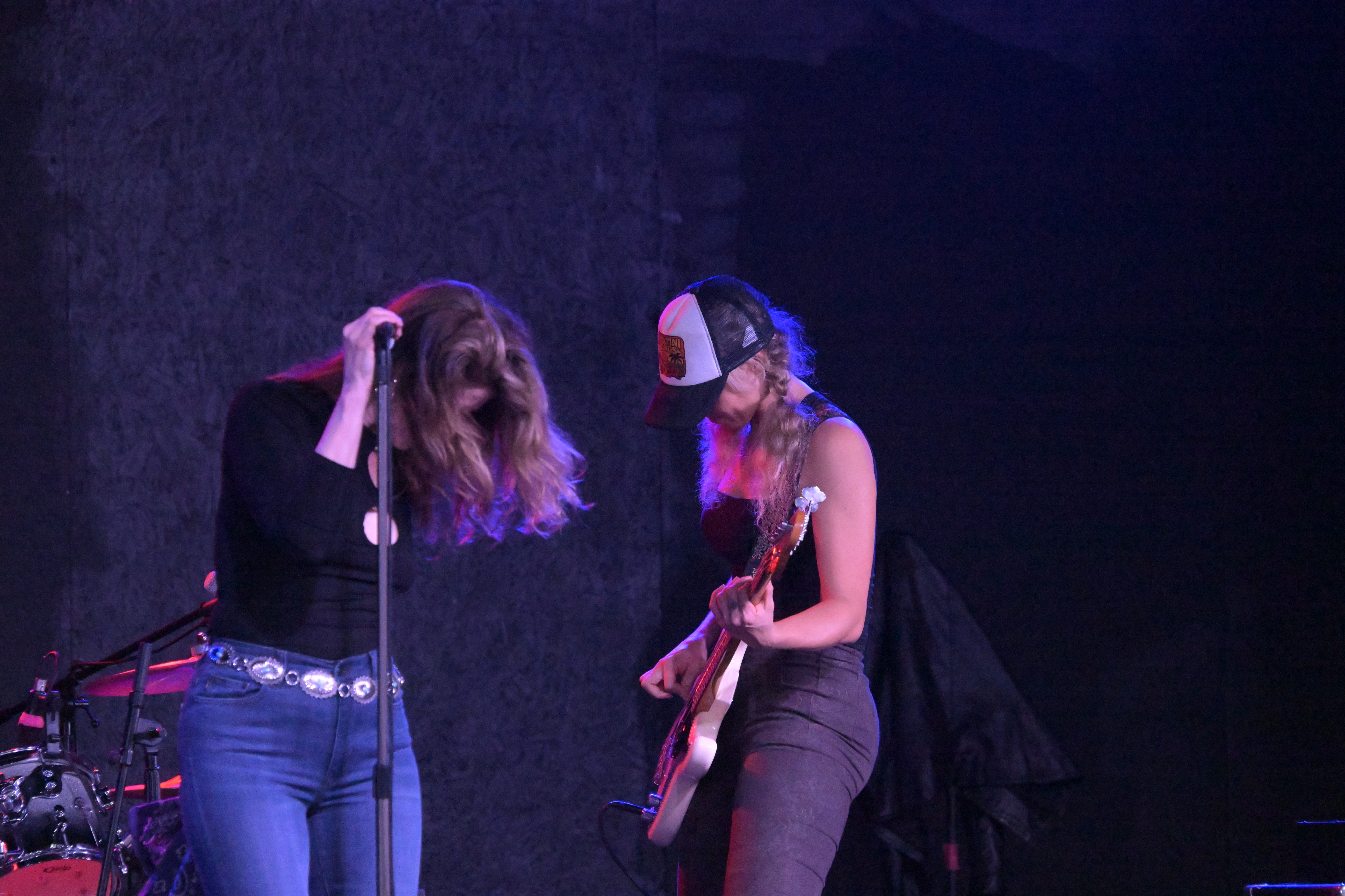Two women on stage, one with long hair and the other wearing a mask, appear to be performing with a guitar.