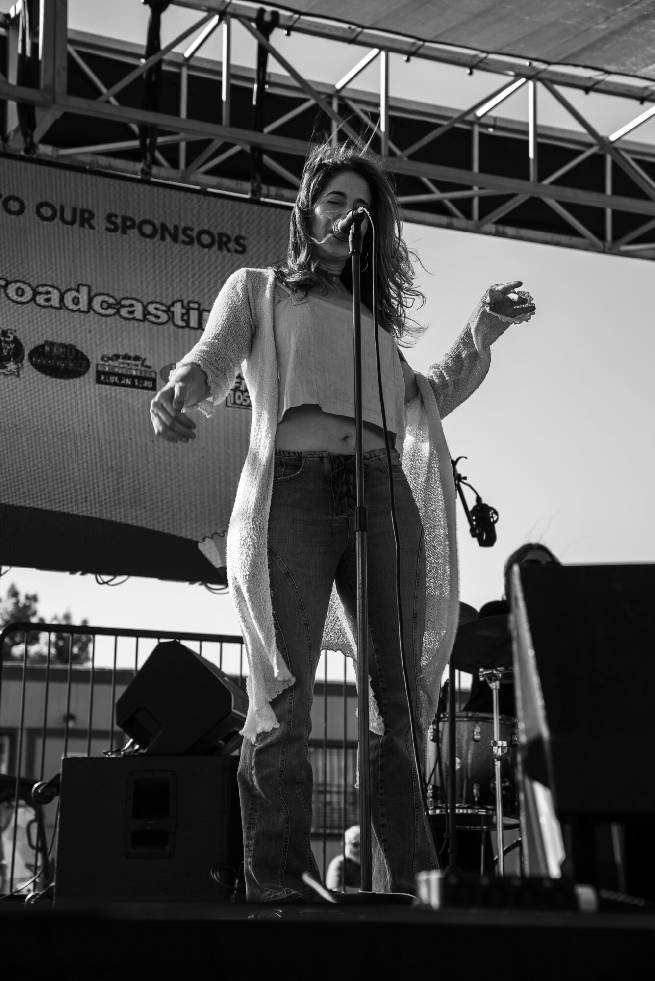 Woman speaking into microphone on stage with a backdrop and stage equipment, outdoors, in black and white.