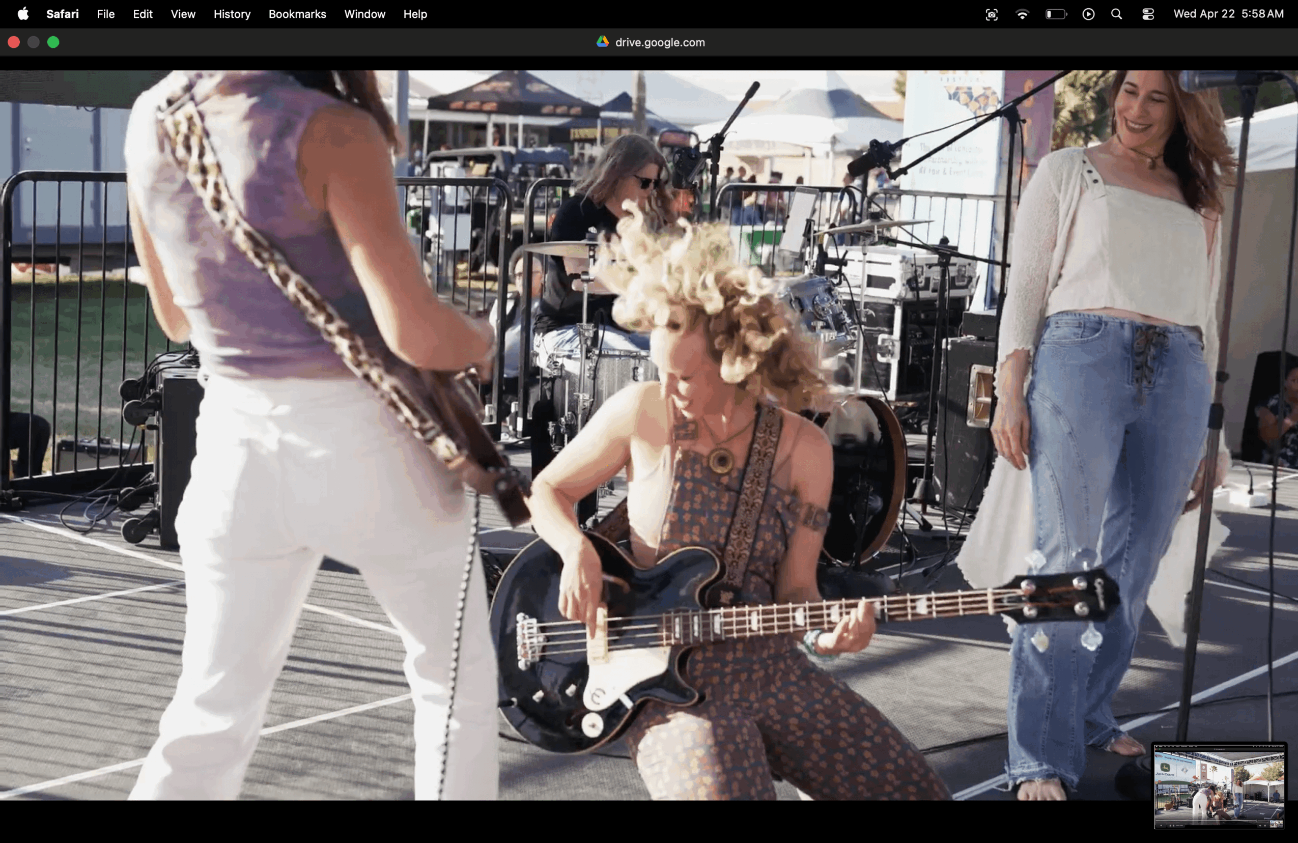 Three women outdoors, one playing guitar, others standing nearby, with musical equipment and a crowd in the background.