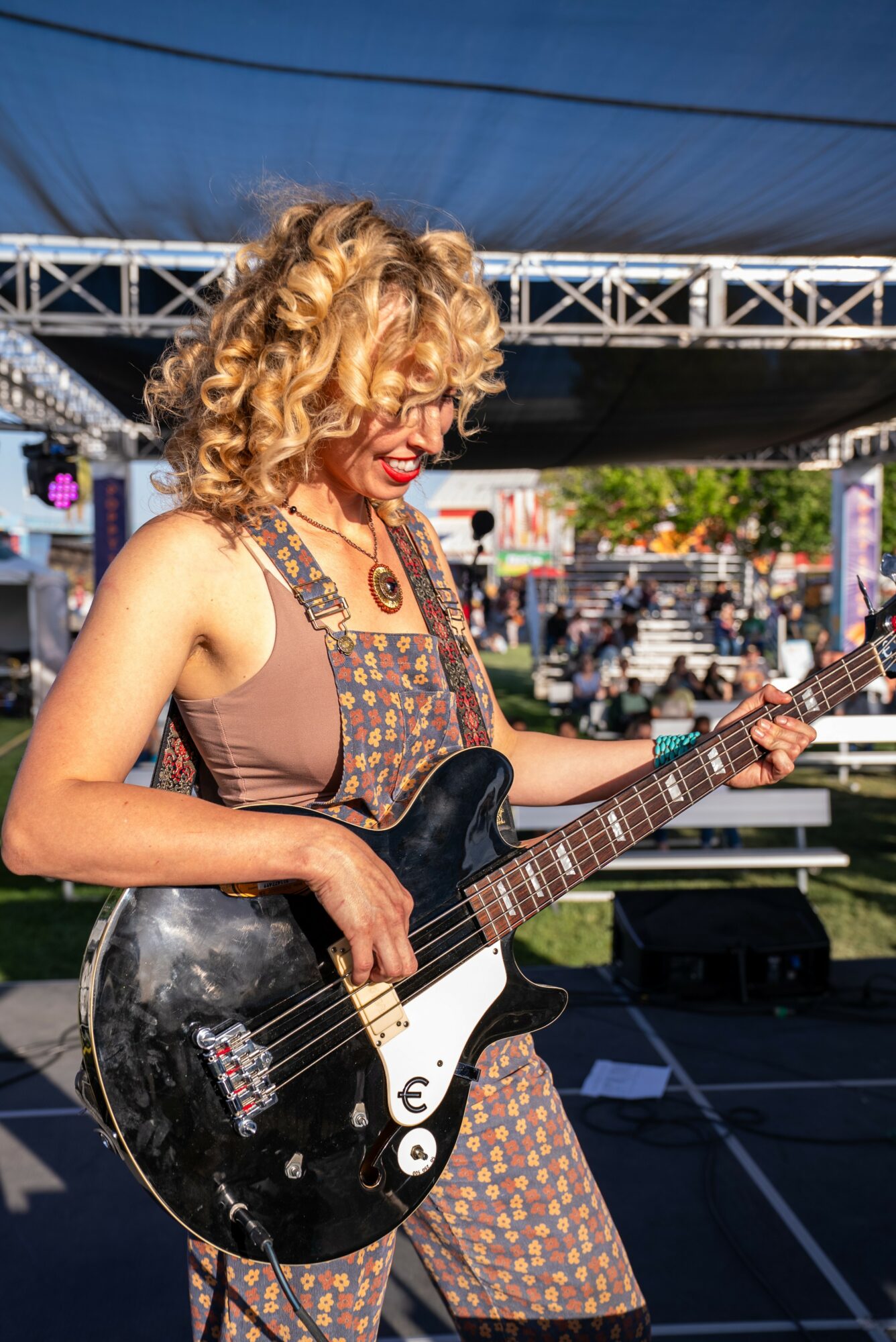 Woman with curly blonde hair playing a black electric guitar outdoors on stage, smiling, with a bridge and audience in background.