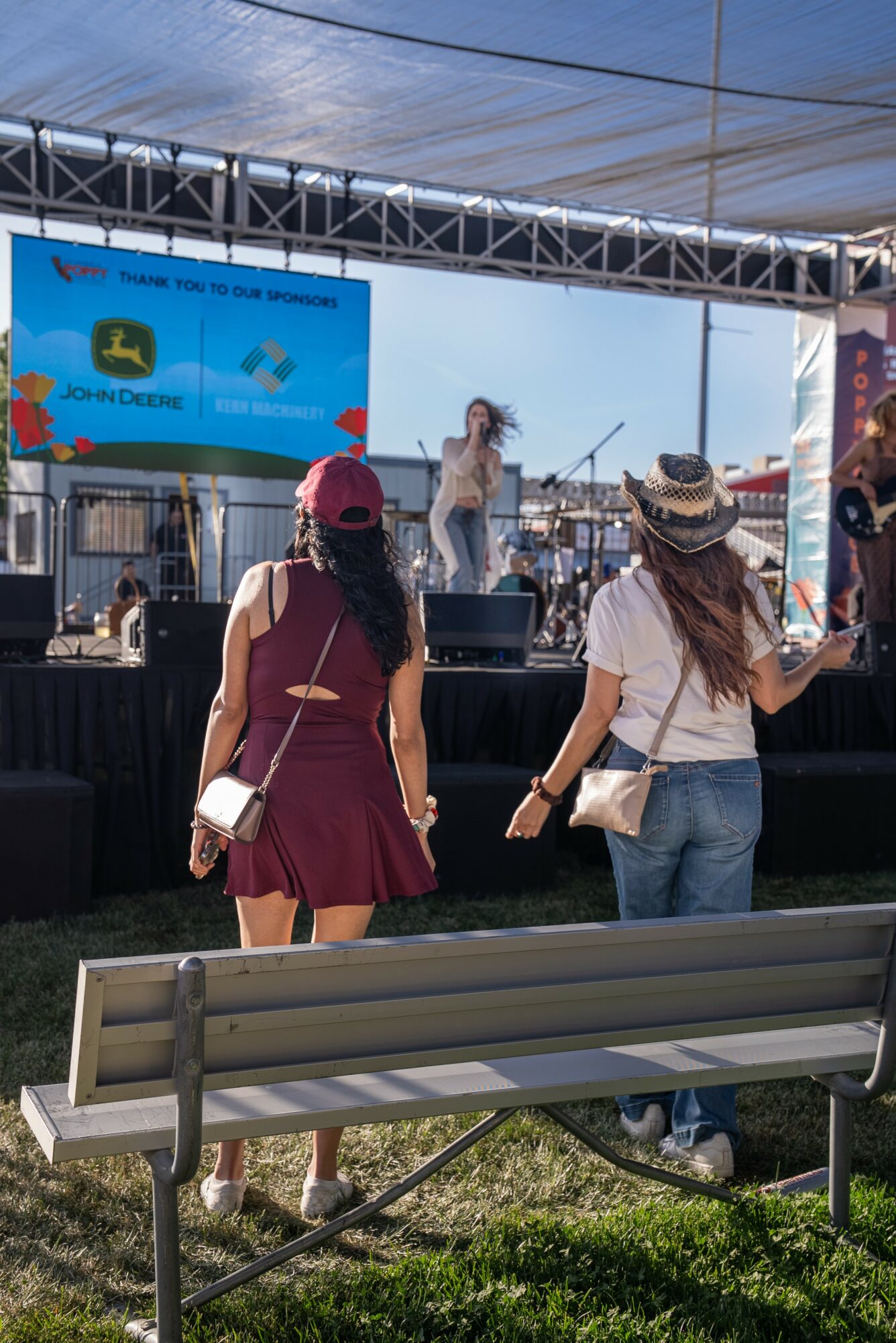 Two women watch a live music performance on an outdoor stage with a singer, under a large canopy, with a blue sky.