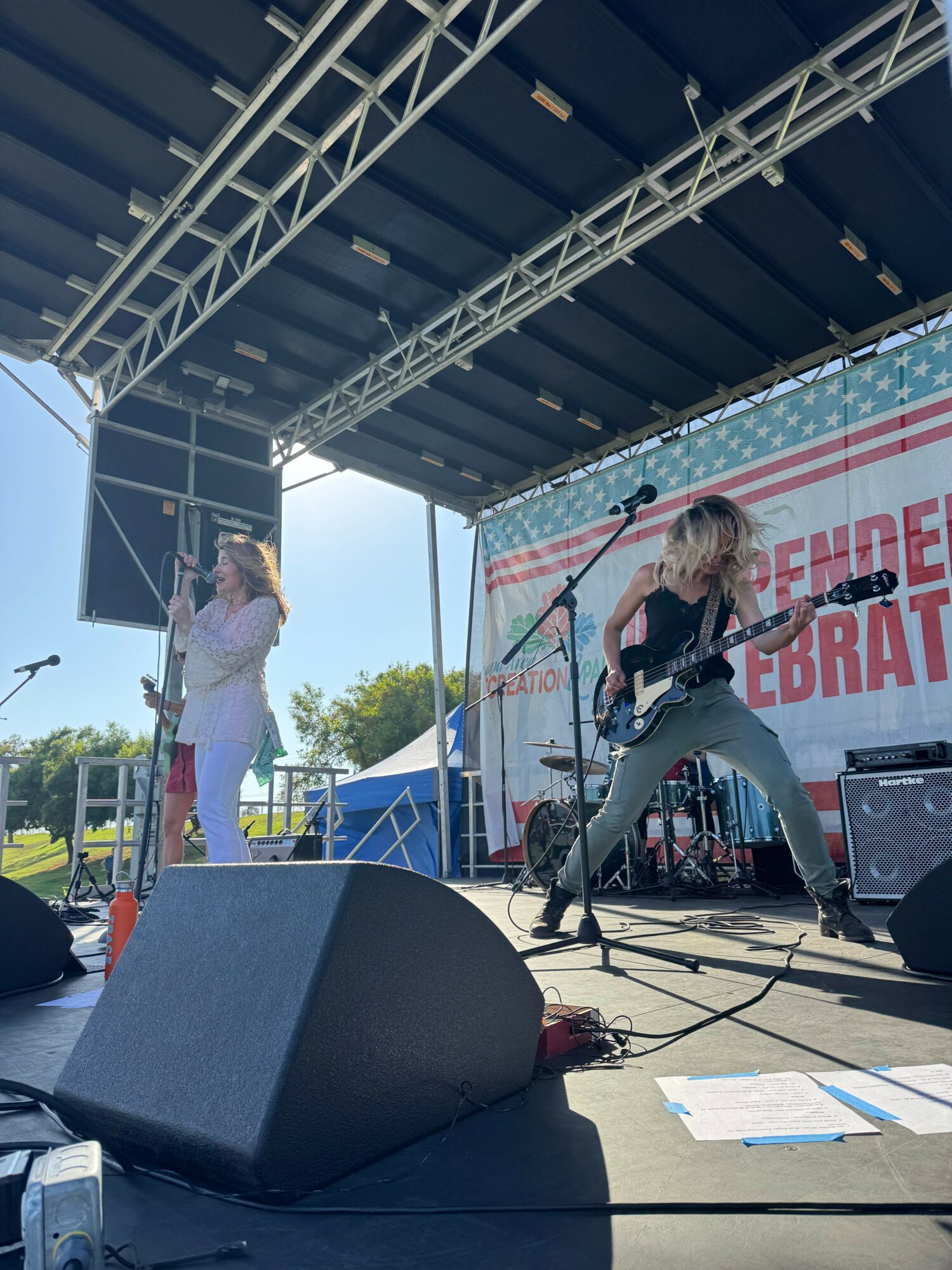 Two performers on stage, one singing into a microphone and the other playing guitar, with a large banner in the background.