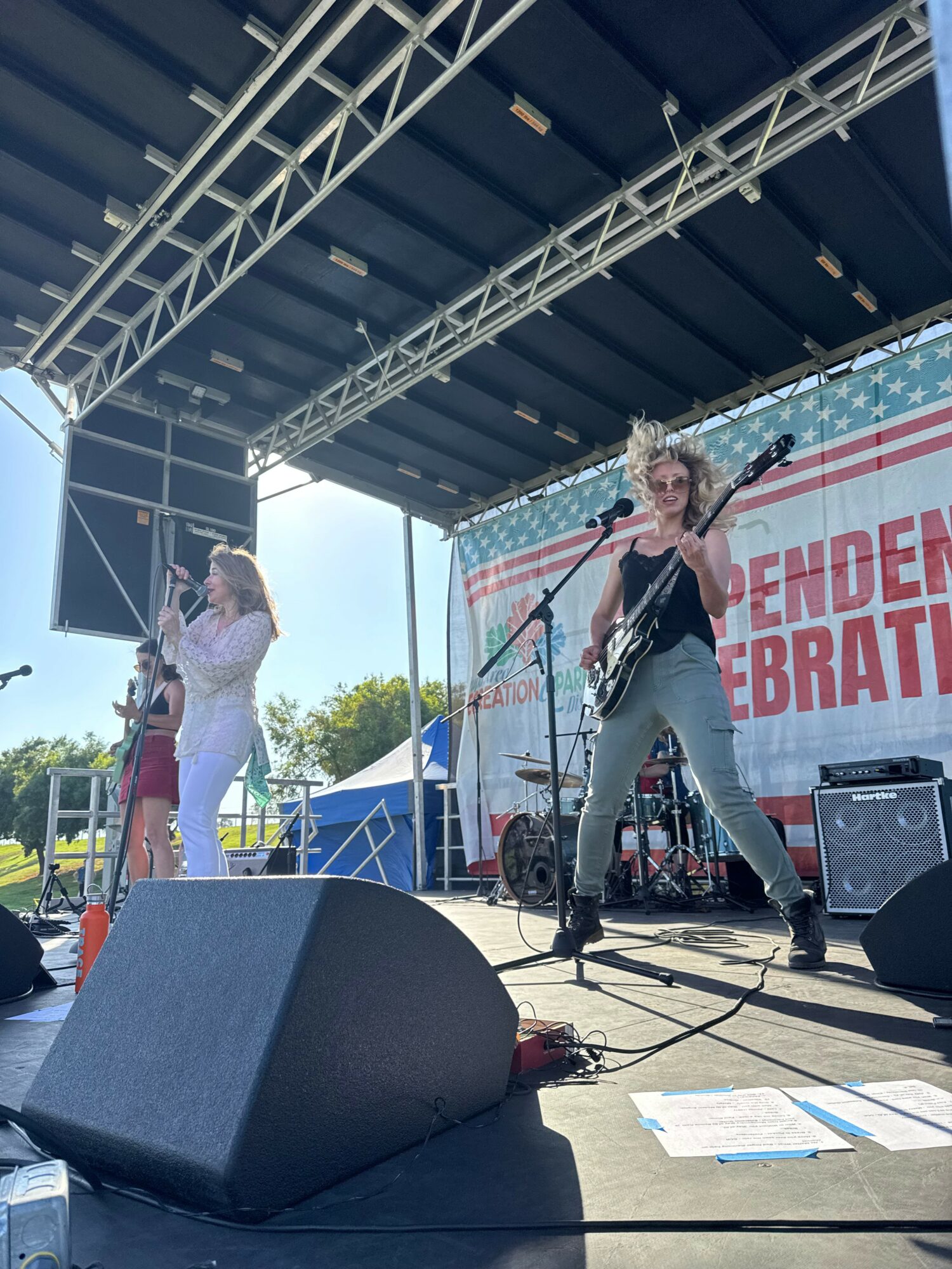 Three women perform on an outdoor stage with musical instruments, under a large canopy, with a banner in the background.