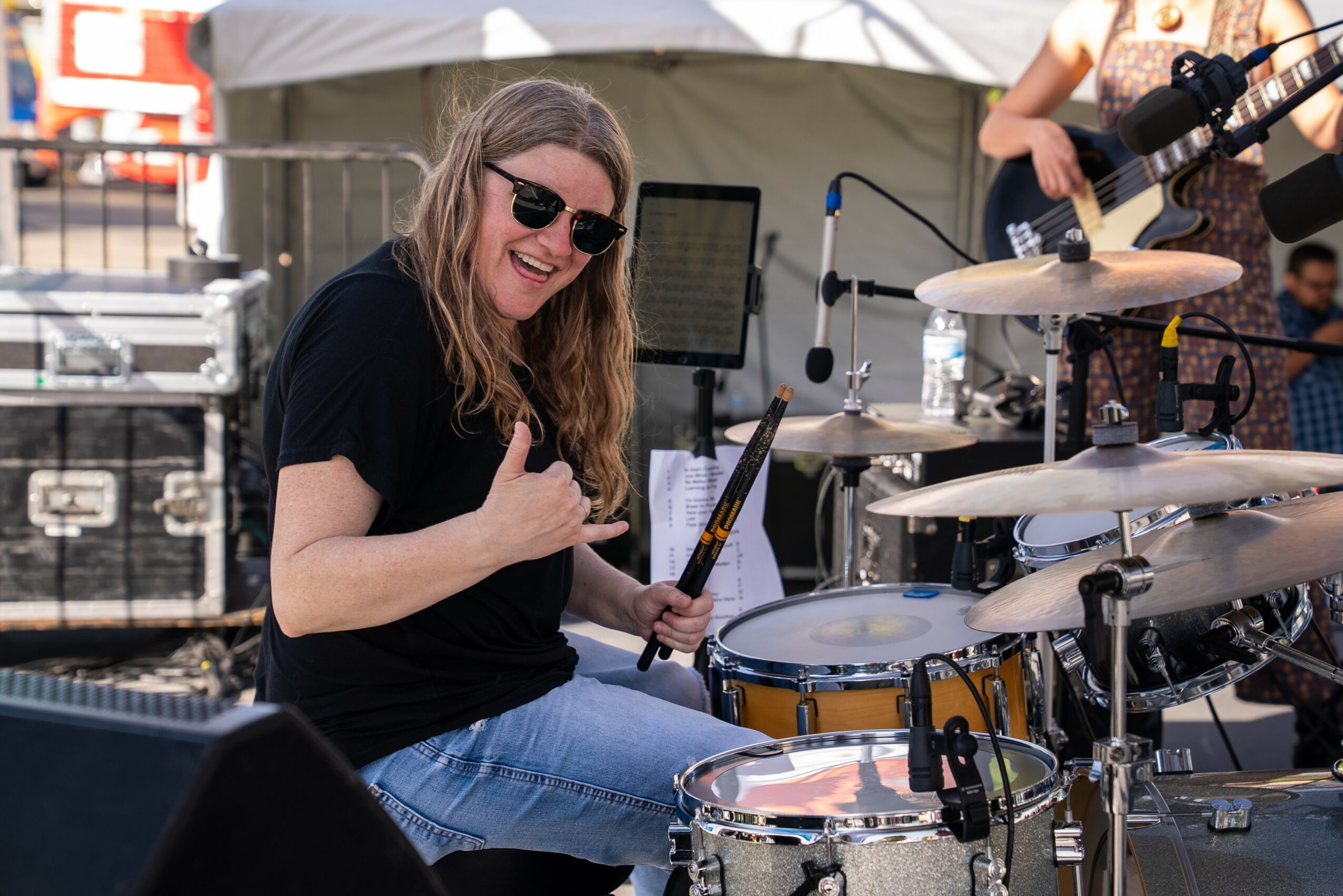 Woman playing drums outdoors, smiling, wearing sunglasses and a black shirt, with a tent and equipment in background.