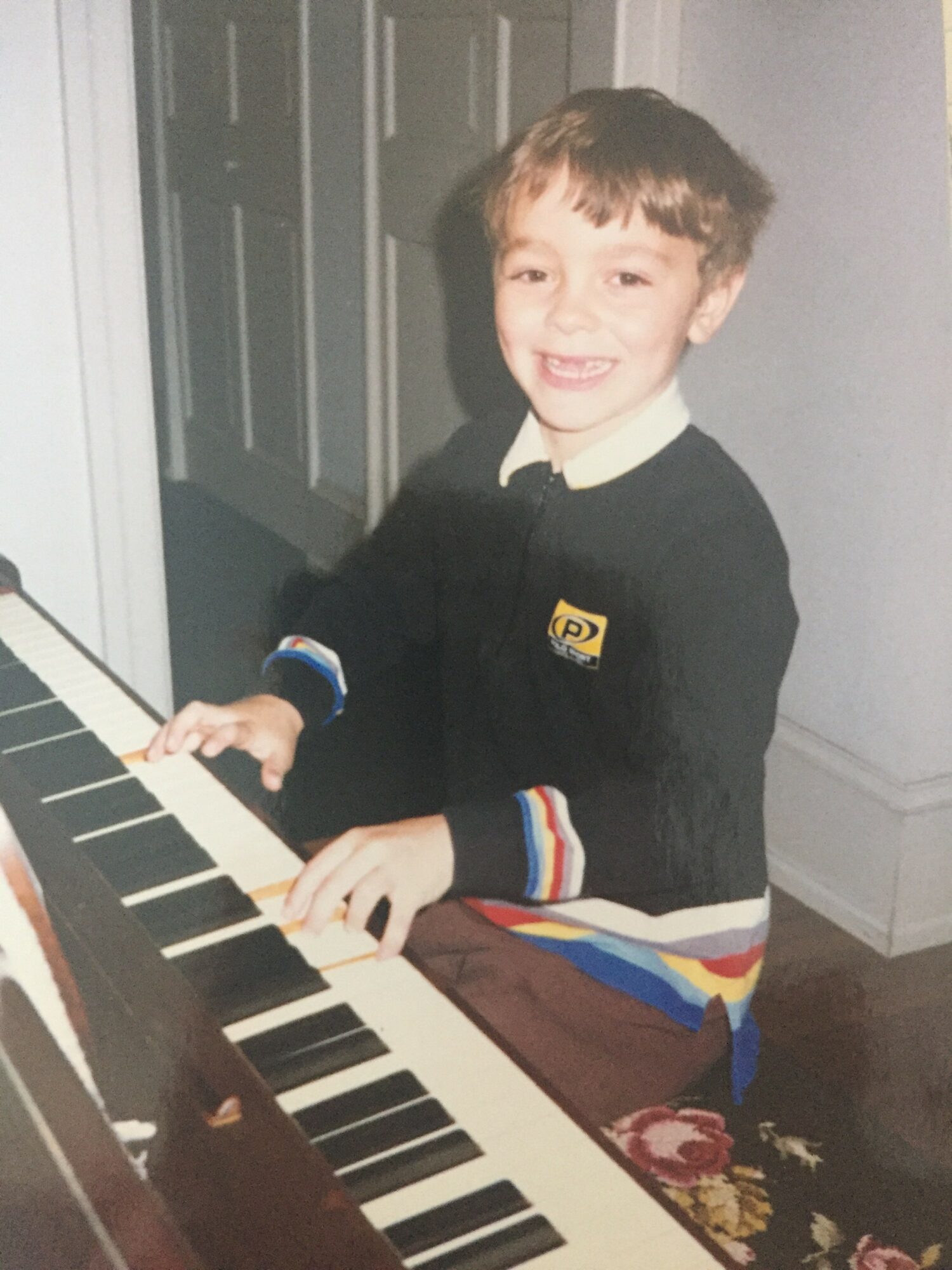 Young boy playing piano, smiling, wearing a black sweater with colorful striped cuffs, in a room with white walls and a door.