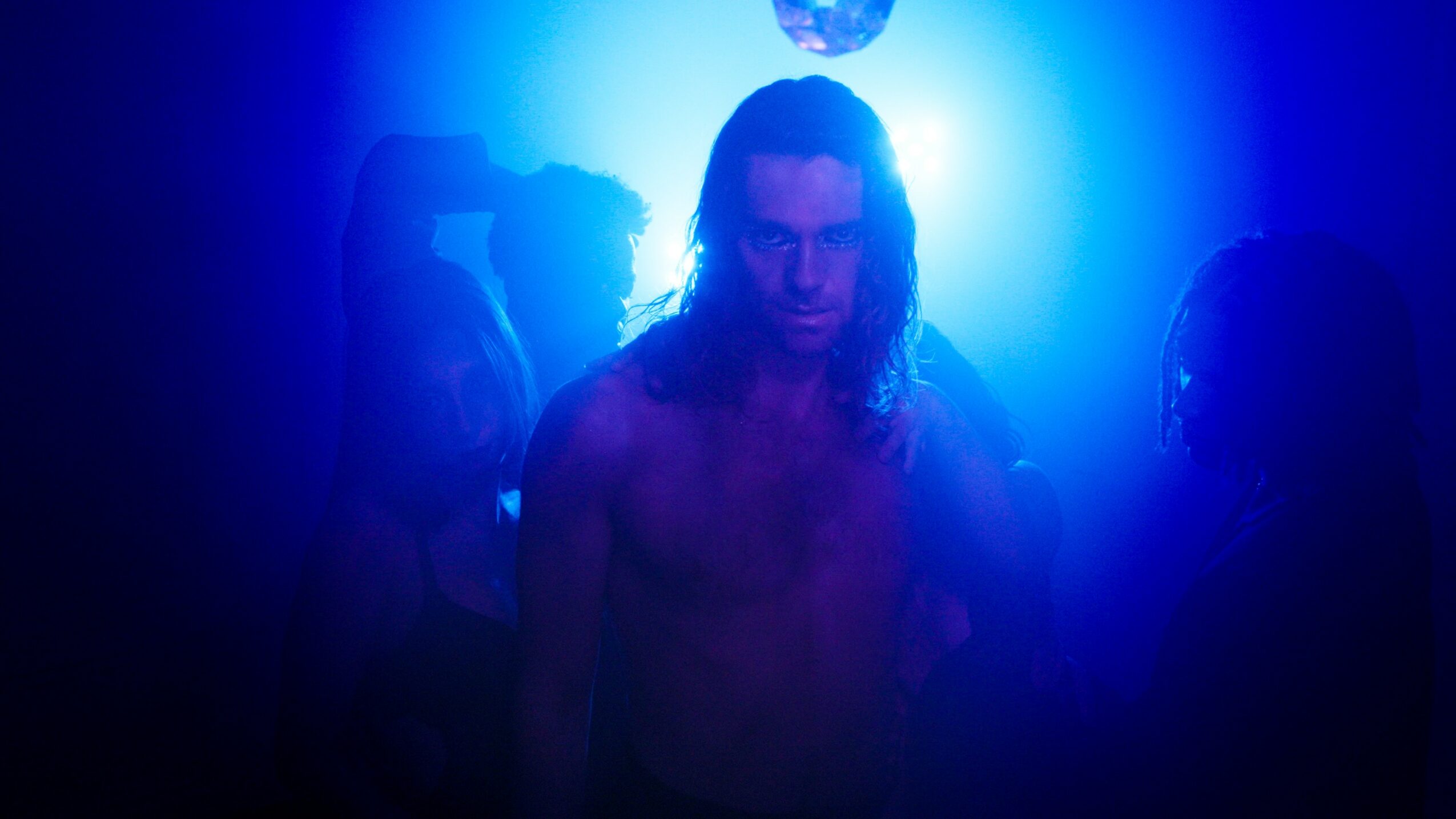 Person with long hair in a dark, blue-lit environment, surrounded by silhouettes of other people.