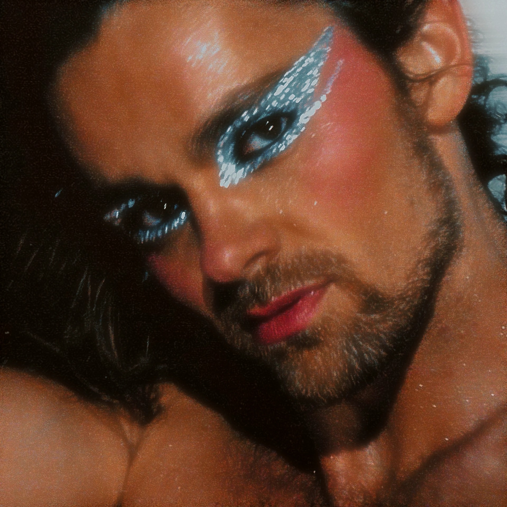 Close-up of a person with elaborate blue and white eye makeup, dark hair, and a beard, looking at the camera.