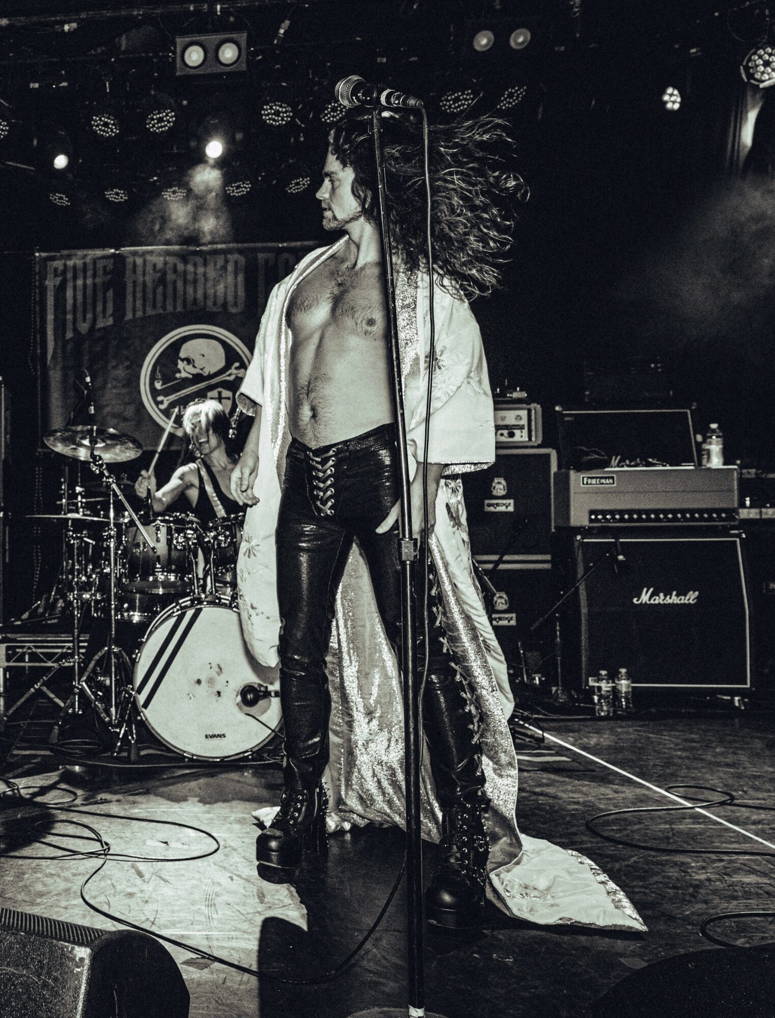 Performer with long hair, shirt open, leather pants, on stage with musical equipment and drum set behind.