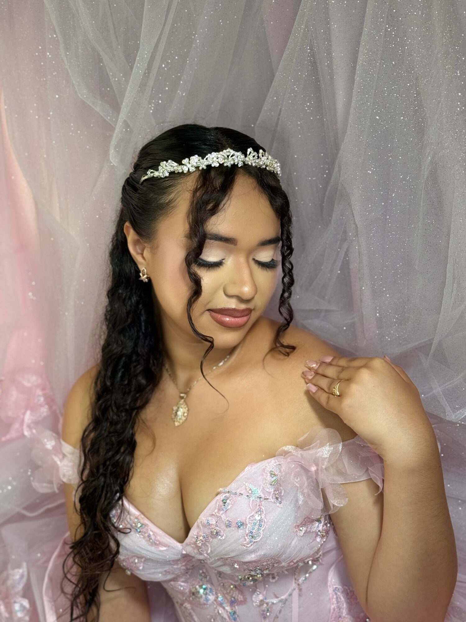 Young woman with long dark hair wearing a floral dress and a tiara, standing in front of sheer fabric backdrop.