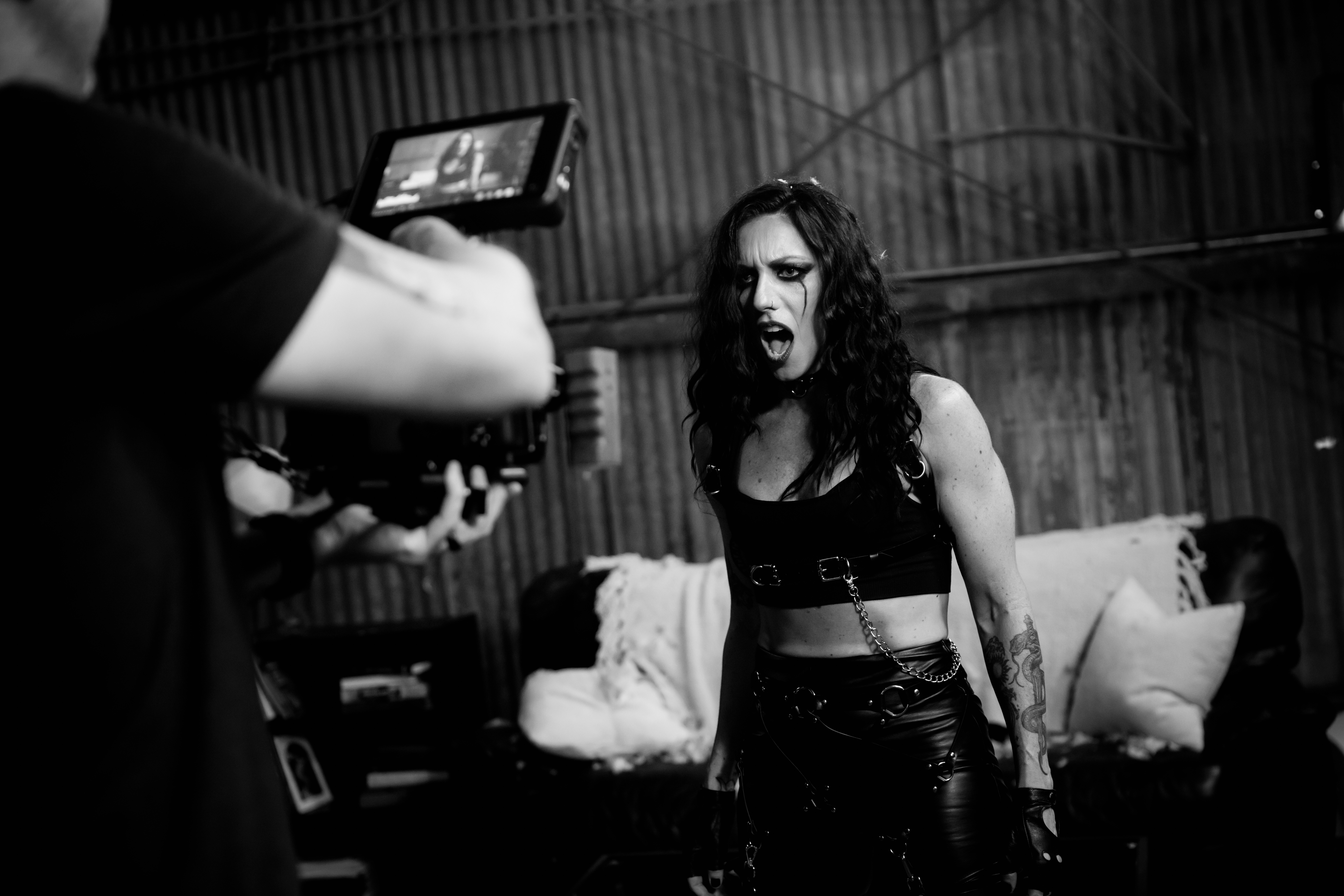 Woman with long hair and tattoos standing indoors, with a person holding a camera in the foreground, black and white photo.