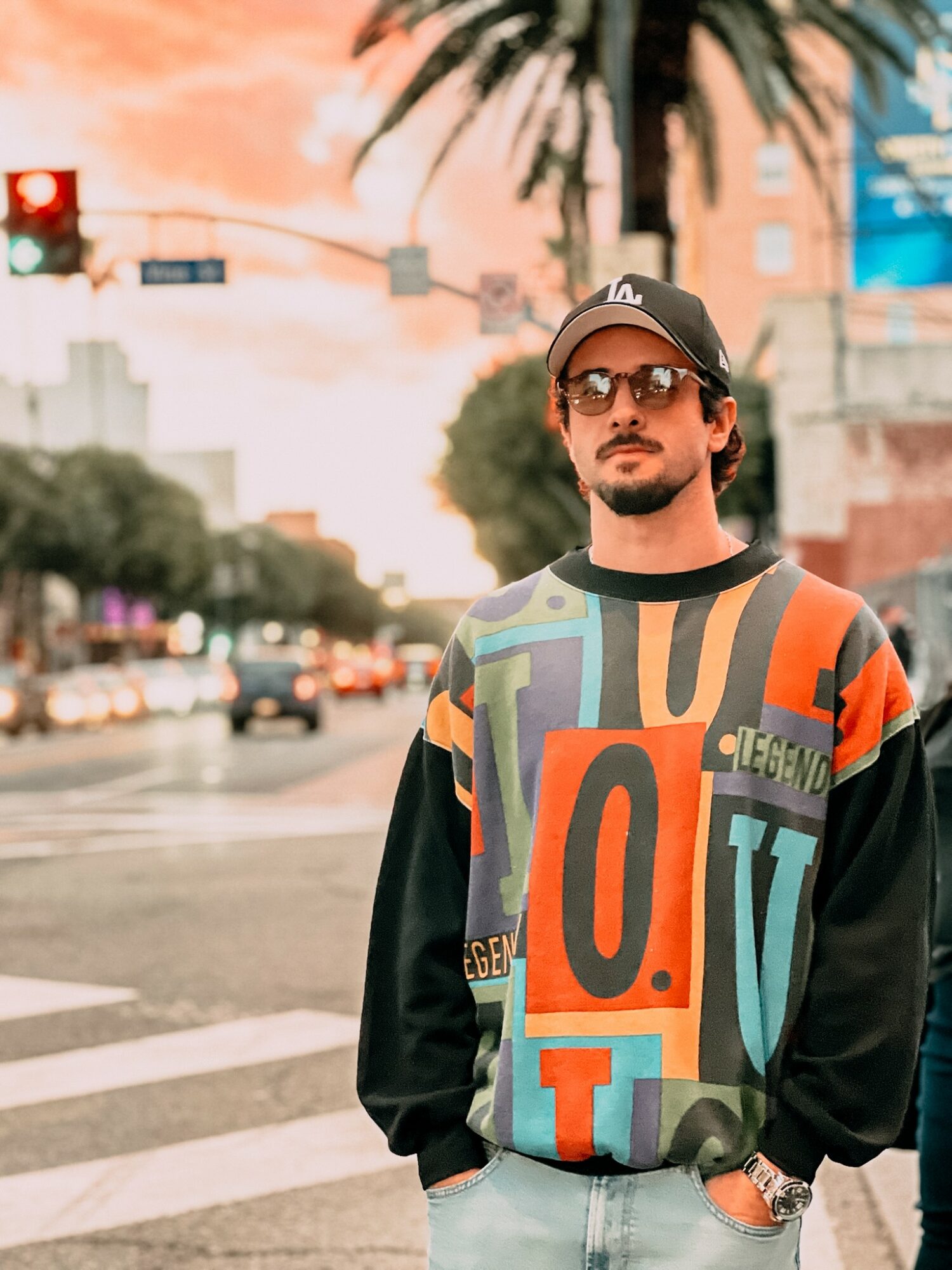 Man with sunglasses and a cap standing on a city street at sunset, wearing a colorful patterned sweatshirt.
