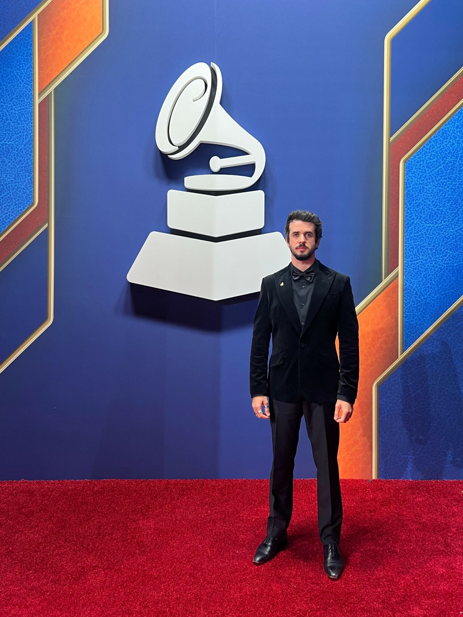 Man in black suit standing on red carpet in front of a blue wall with a large Grammy Award logo.