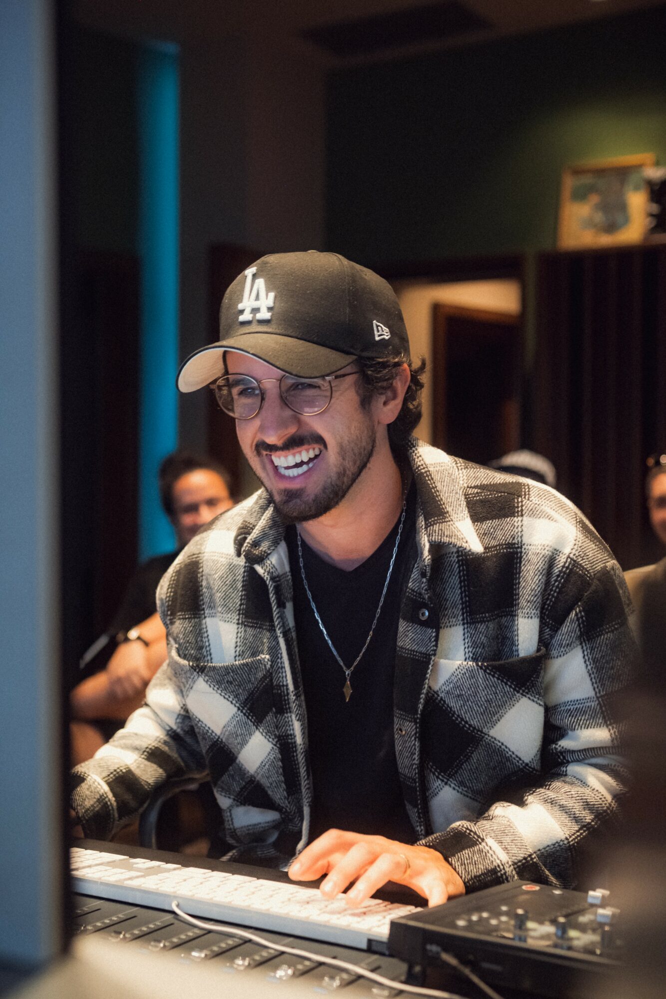 Man with glasses, baseball cap, and plaid shirt smiling while playing keyboard in a room with people.